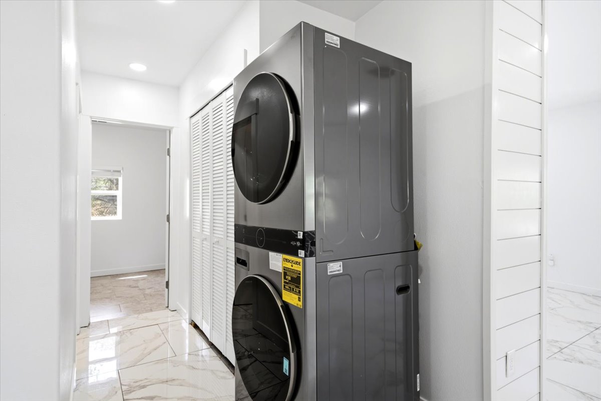 Integrated laundry center with stacked front-load machines, louvered bi-fold doors, recessed ceiling lighting, and polished stone-look tile flooring