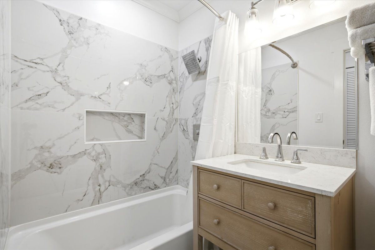 Bathroom featuring a white bathtub, marble-look tiled shower surround with an integrated niche, a light wood-finish vanity with drawer storage, a white countertop with an integrated sink, and a large mirror