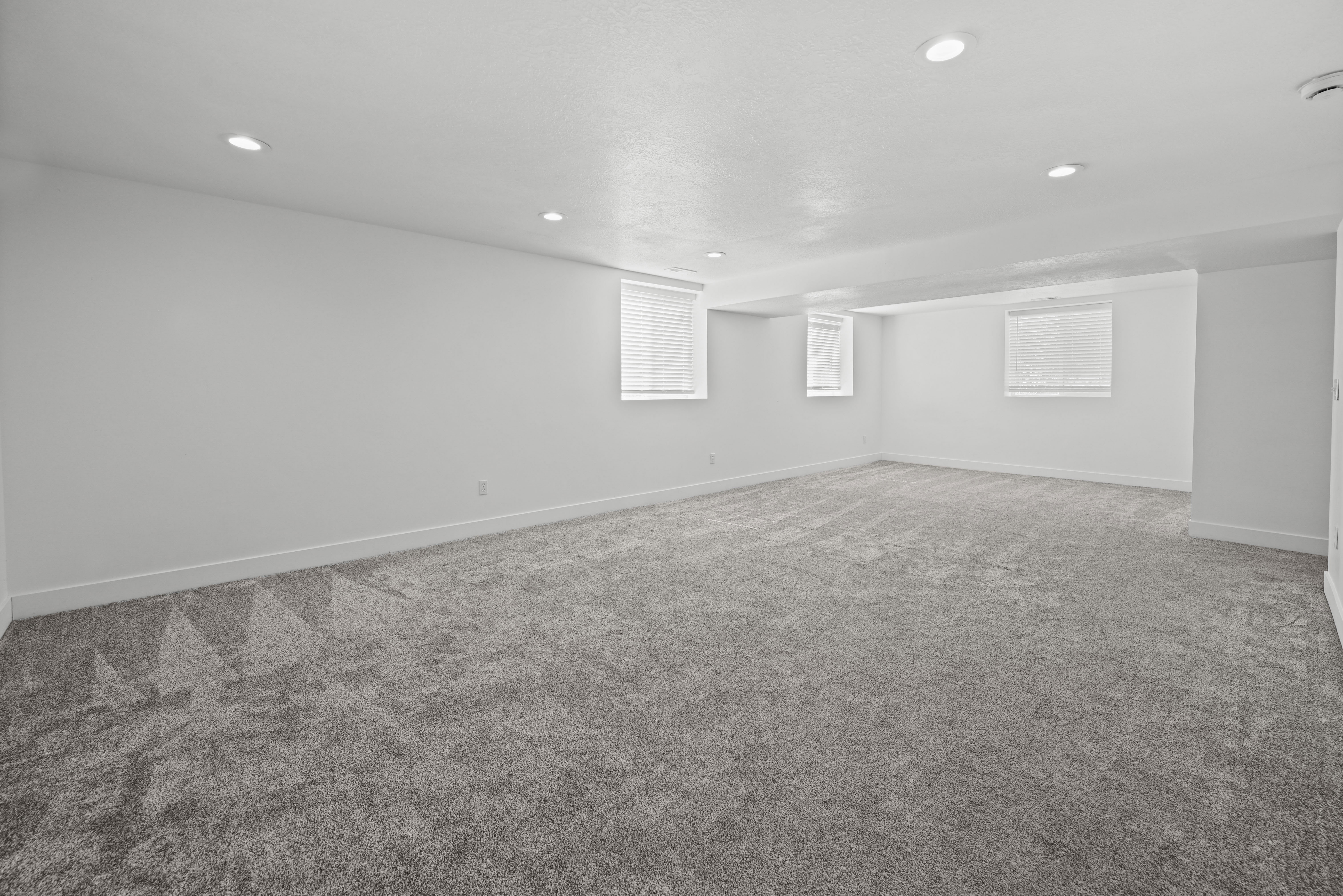 Spacious room featuring new gray carpet, white walls, recessed ceiling lighting, and multiple windows with blinds