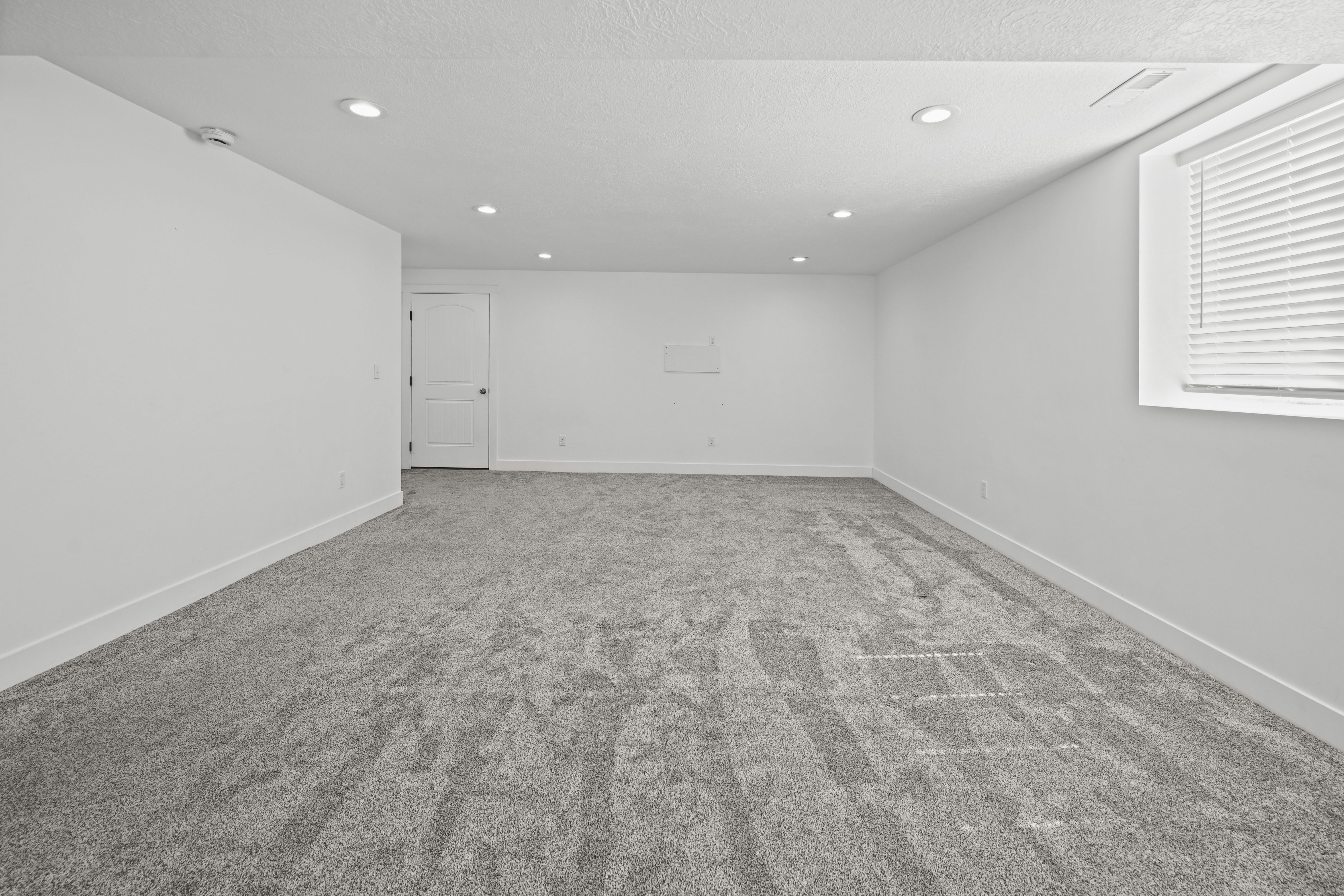 Spacious room featuring gray carpeting, white walls, recessed ceiling lights, a white interior door, and a window with horizontal blinds