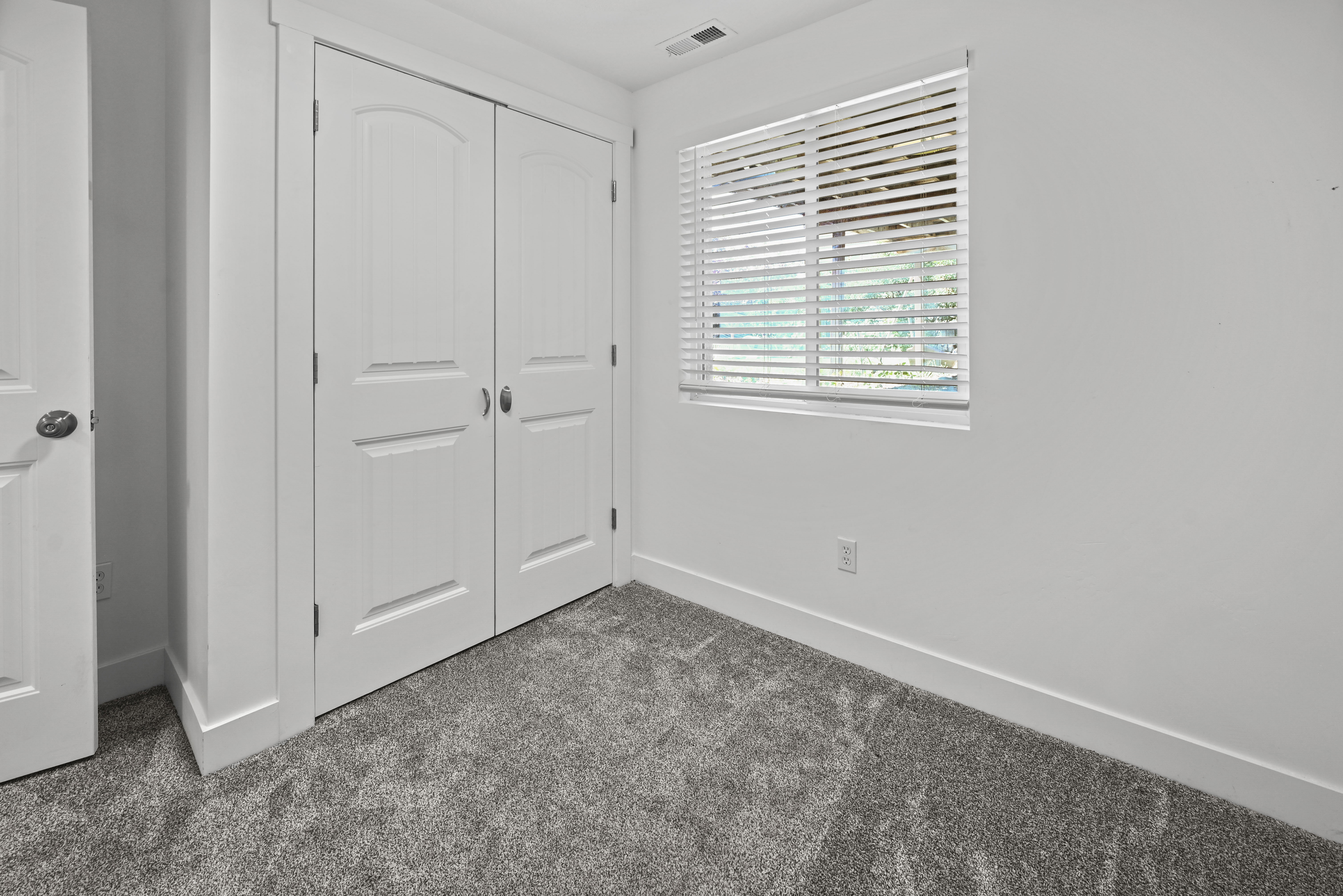 Interior room featuring textured grey carpeting, a double-door closet with arched paneling, and a single window with horizontal blinds