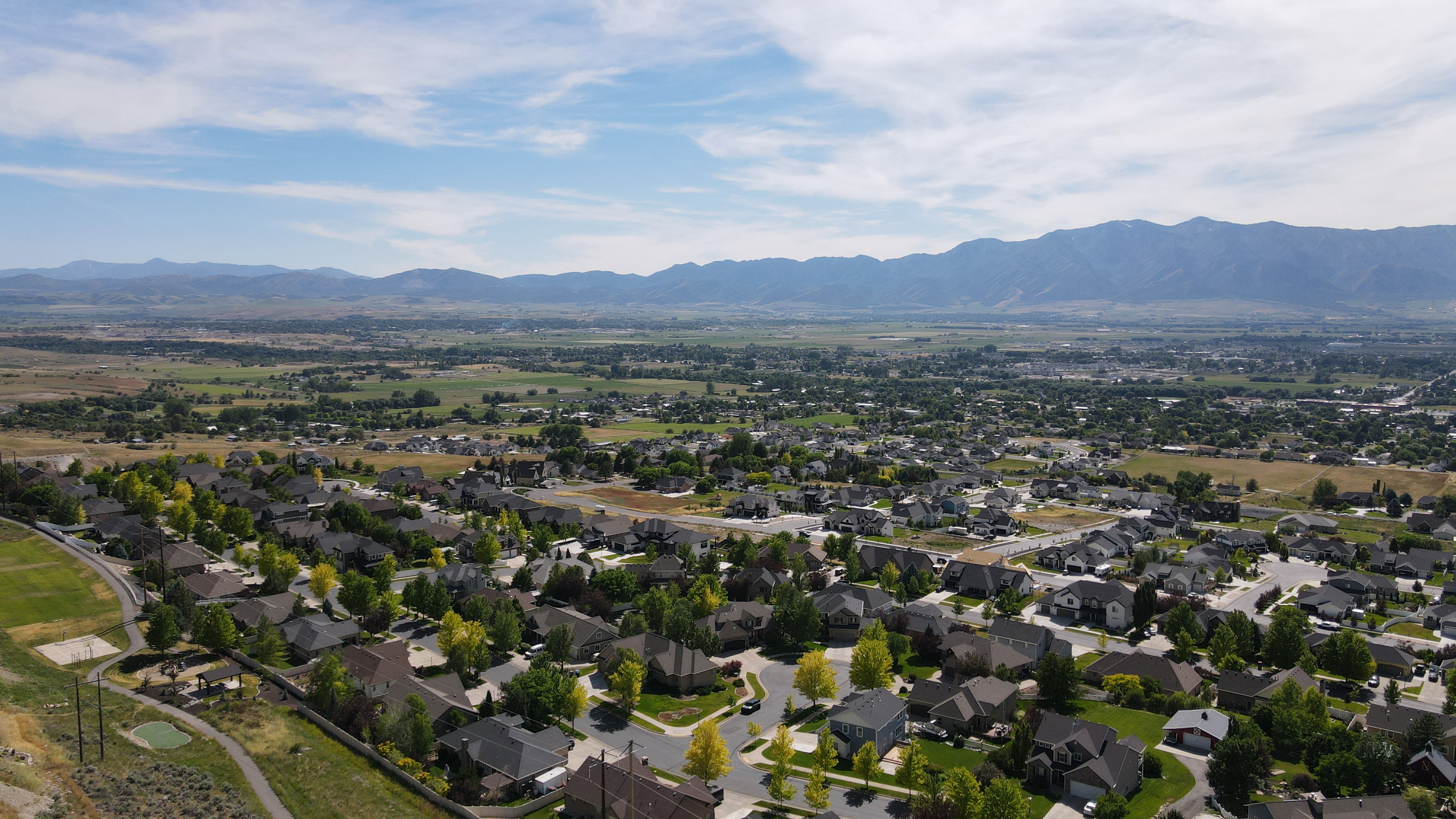 Expansive residential area featuring numerous single-family homes, mature trees, and paved streets, all set against a backdrop of distant mountains and open fields under a blue sky