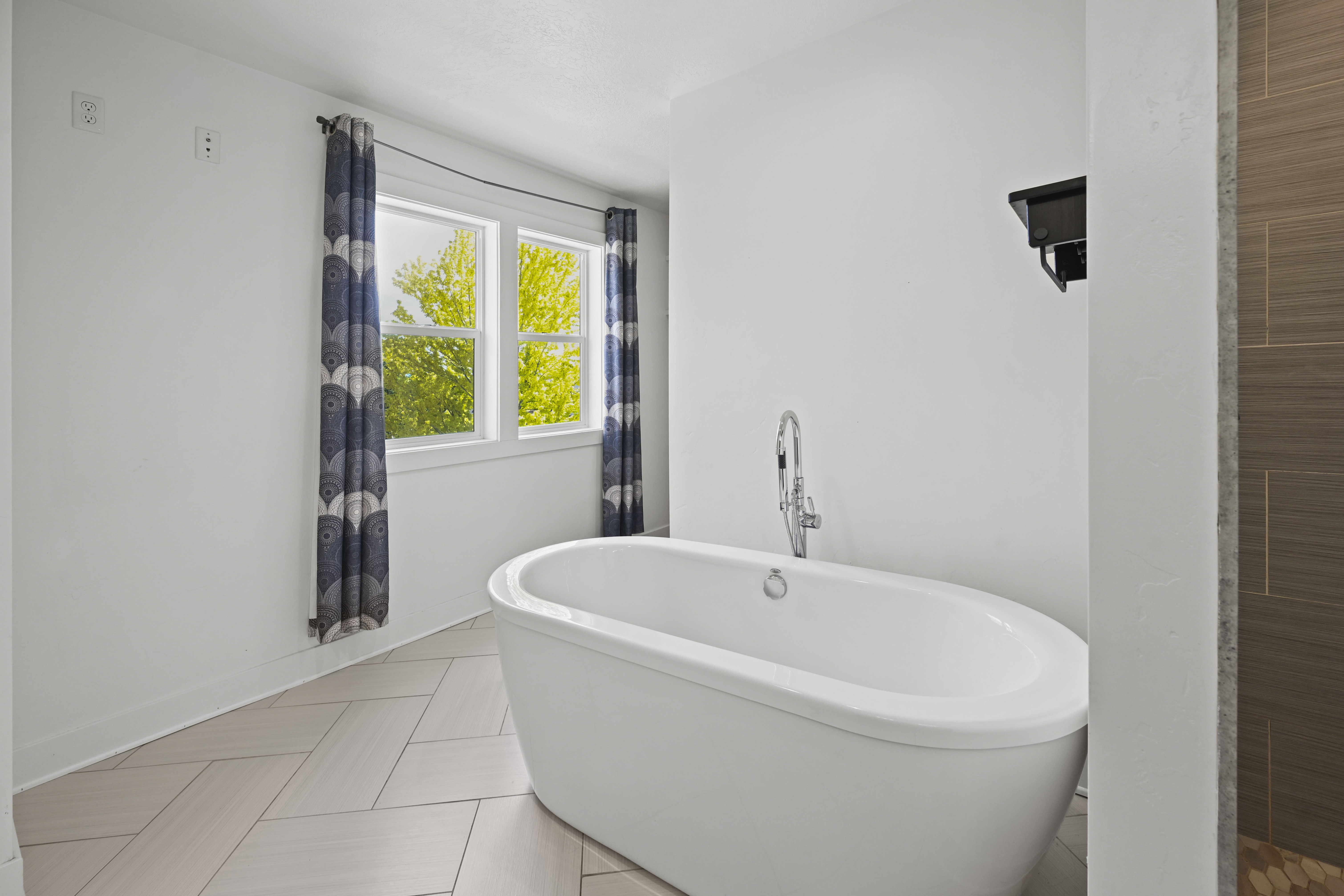 Freestanding oval soaking tub featuring a floor-mounted chrome faucet, set against a backdrop of bright windows with patterned curtains