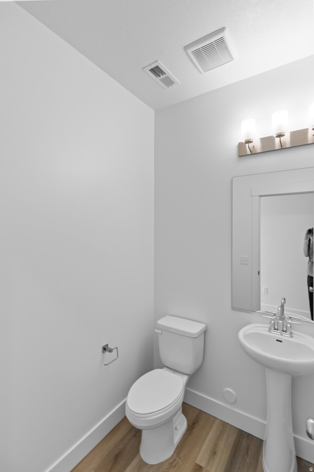Modern bathroom featuring wood-finish flooring, white pedestal sink, chrome faucet, and a wall-mounted mirror with sconce lighting