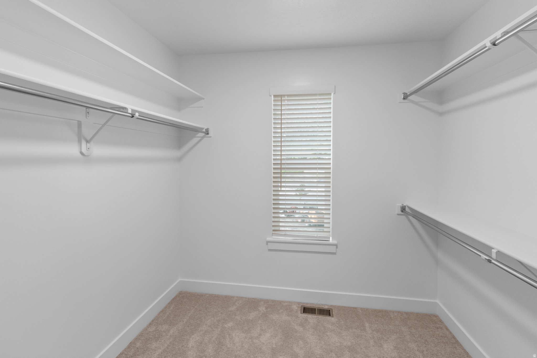 Walk-in closet featuring neutral carpeting, a single window with horizontal blinds, white baseboards, and built-in white shelving with hanging rods