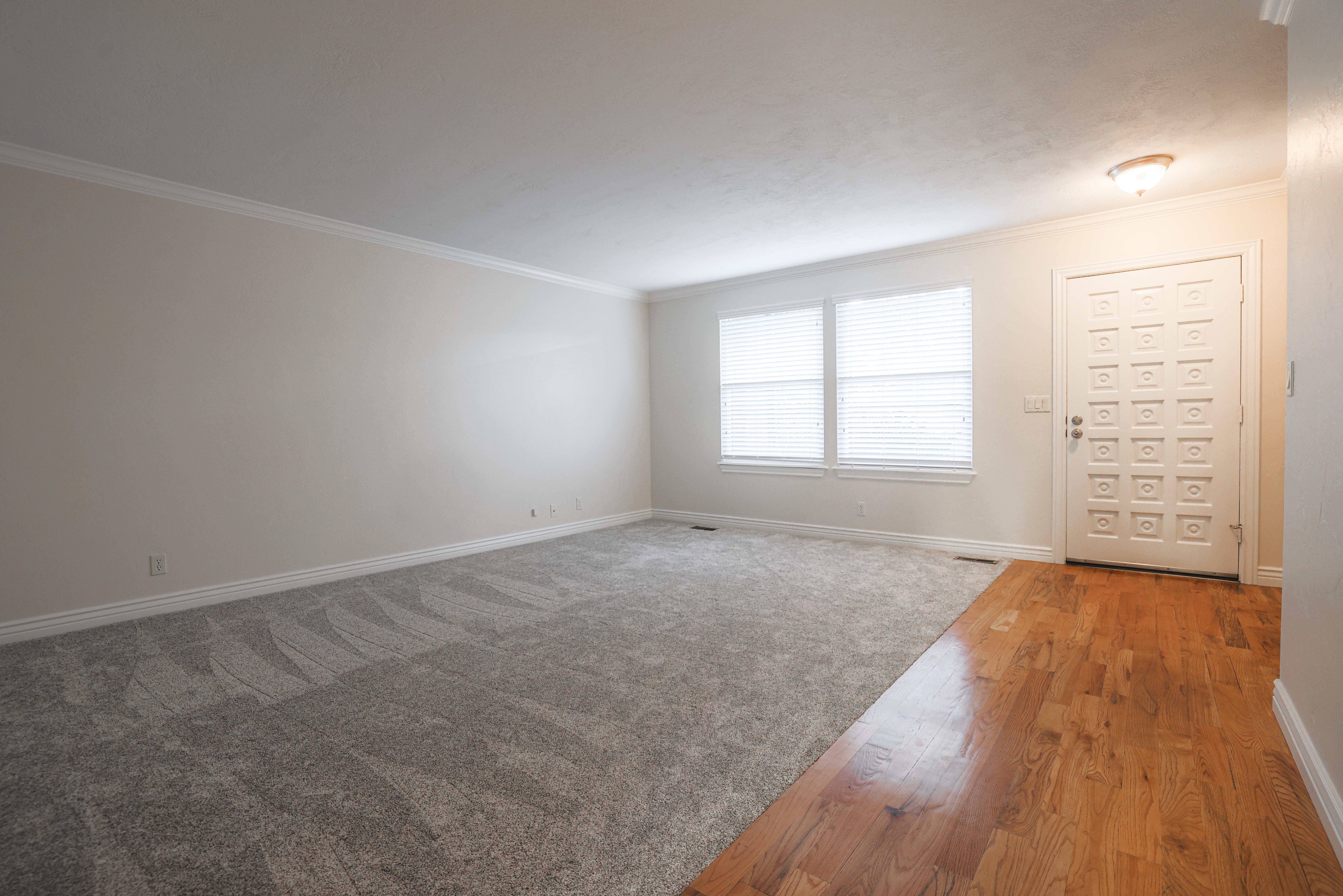 Spacious interior featuring a decorative front entry door, two large windows with blinds, a combination of hardwood and carpet flooring, and crown molding accents
