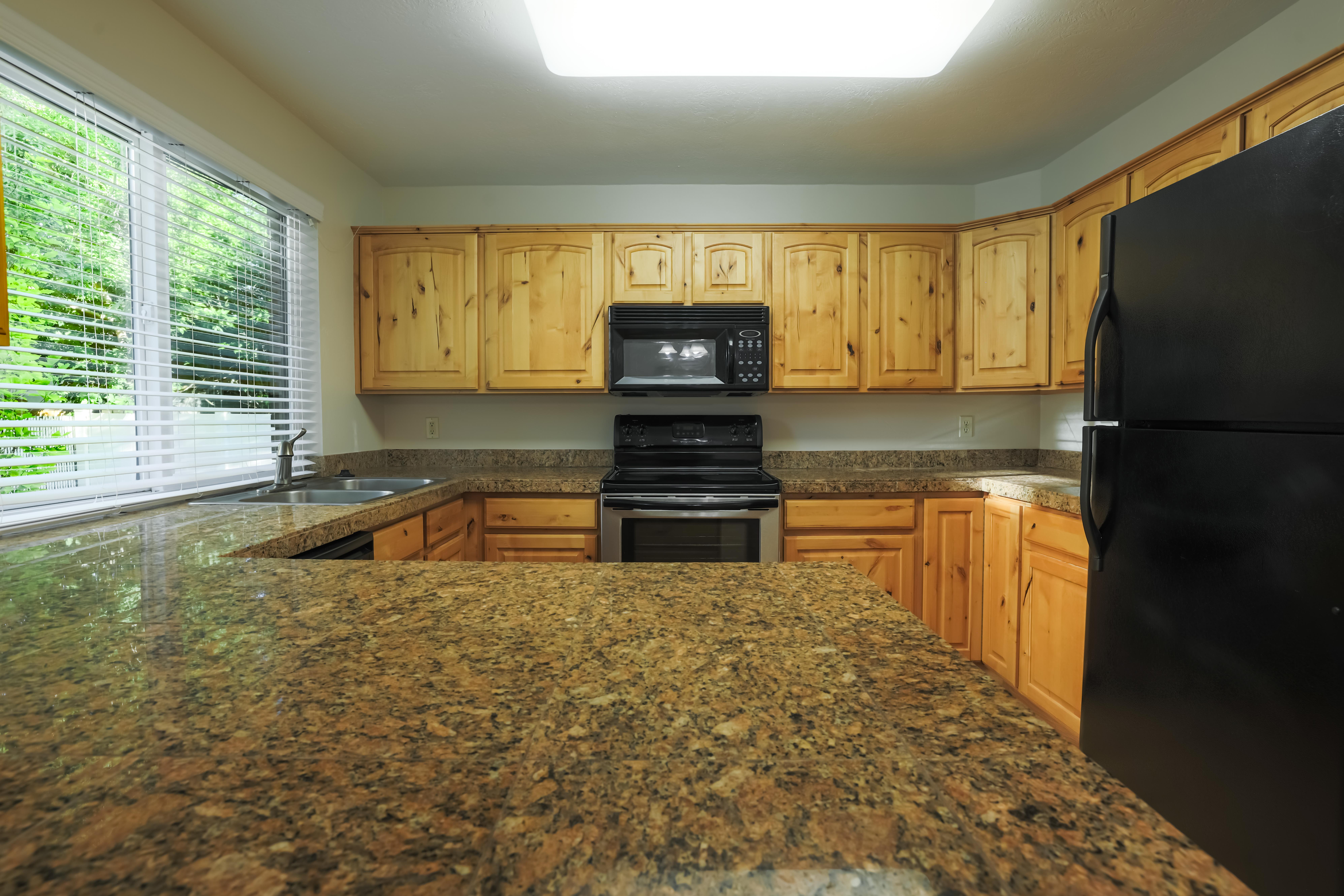 Kitchen featuring extensive stone countertops, natural wood cabinetry, stainless steel sink, black and stainless steel appliances, and a large window