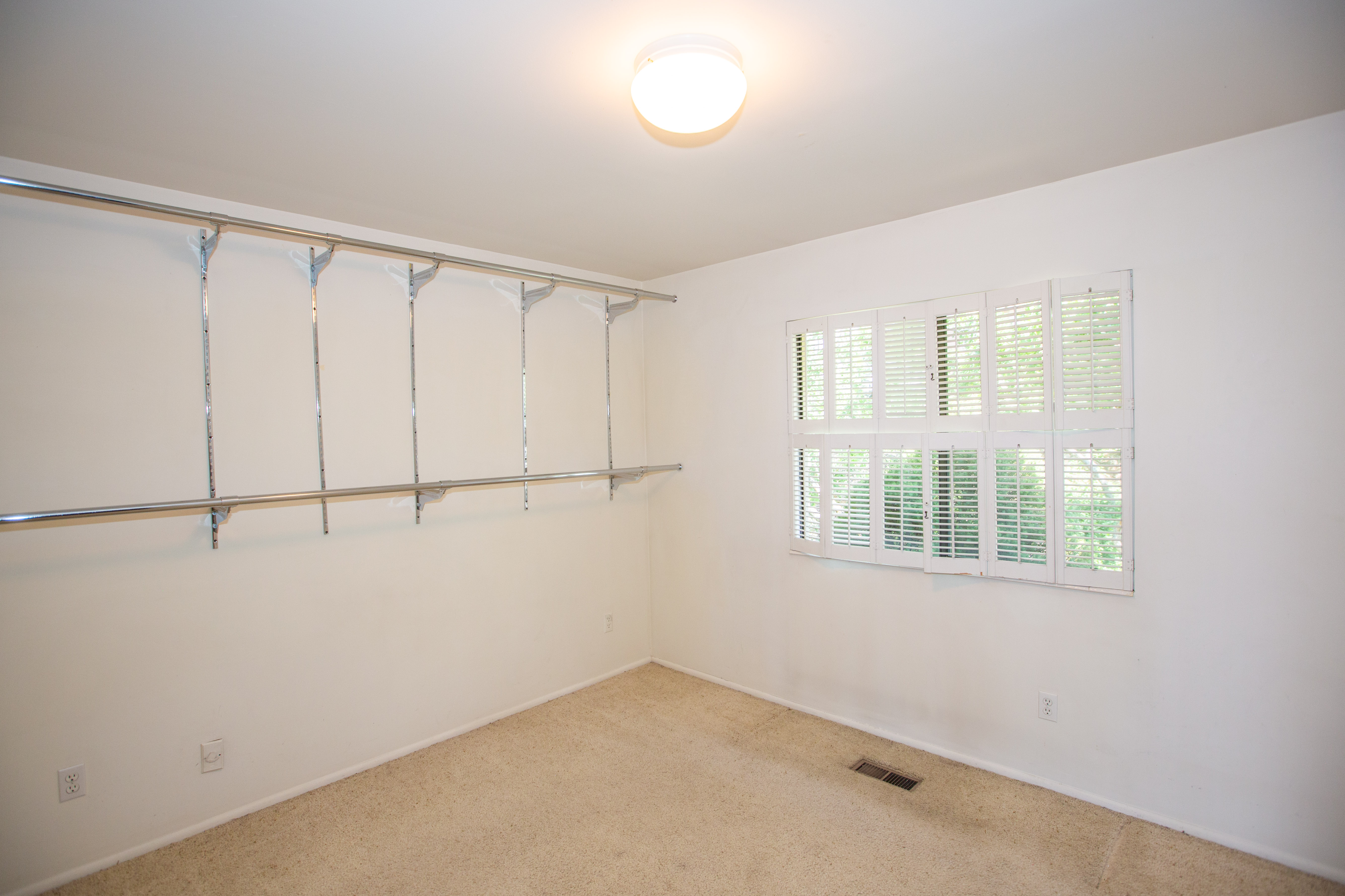 Bedroom featuring wall-mounted shelving and hanging rods, a window with white louvered shutters, carpeted flooring, and a flush-mount ceiling light fixture