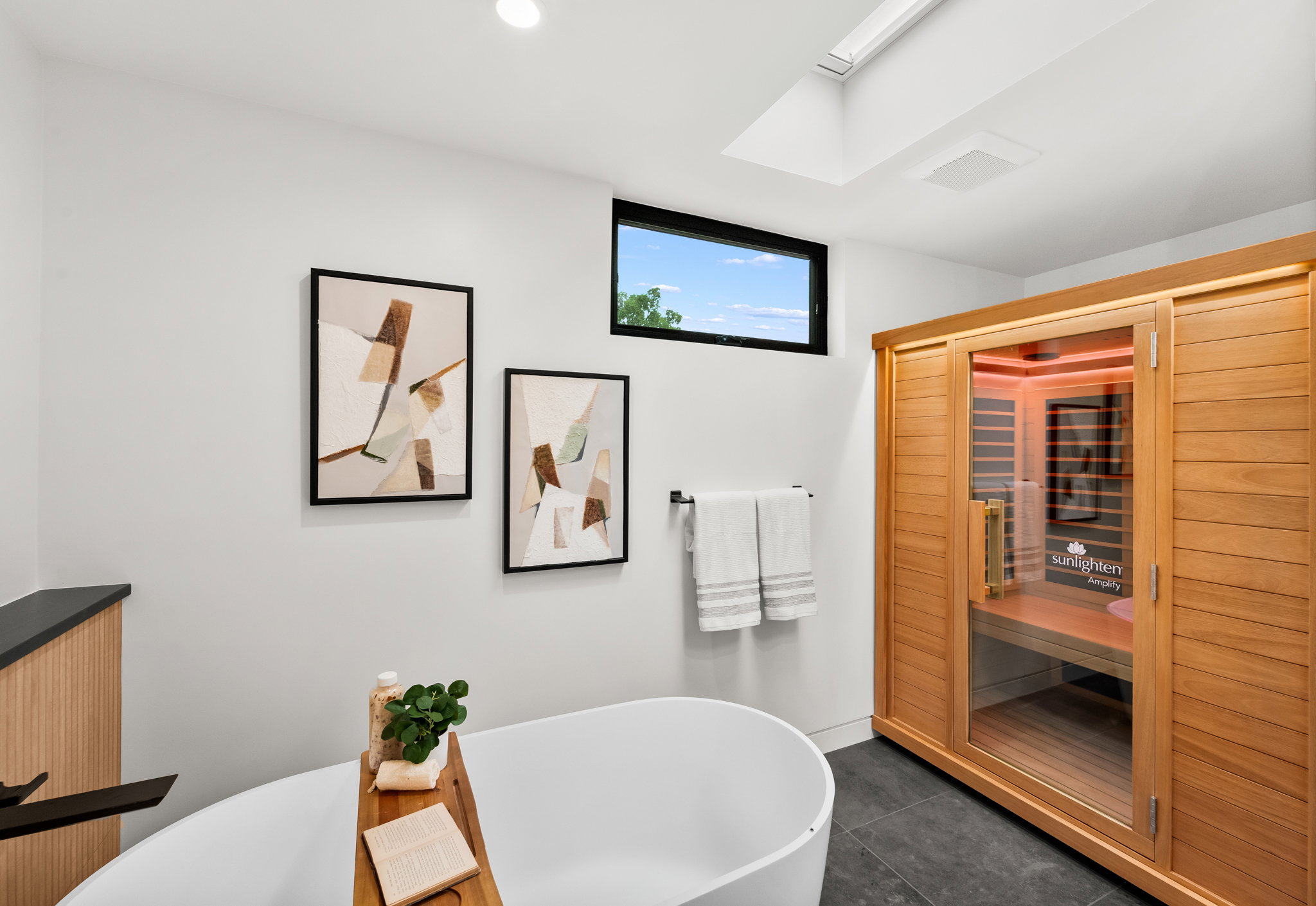 Modern bathroom featuring a freestanding soaking tub, wood-finish sauna with glass door, dark tile flooring, and a skylight