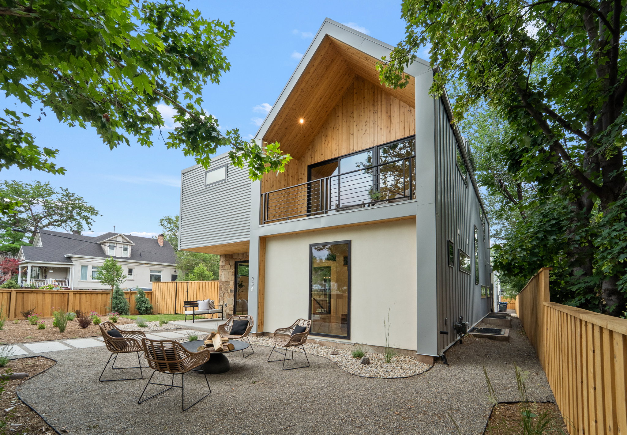 Contemporary architectural design featuring a prominent peaked roof with wood-finish soffit, a second-story balcony with dark metal railing, and a ground-level patio with gravel landscaping