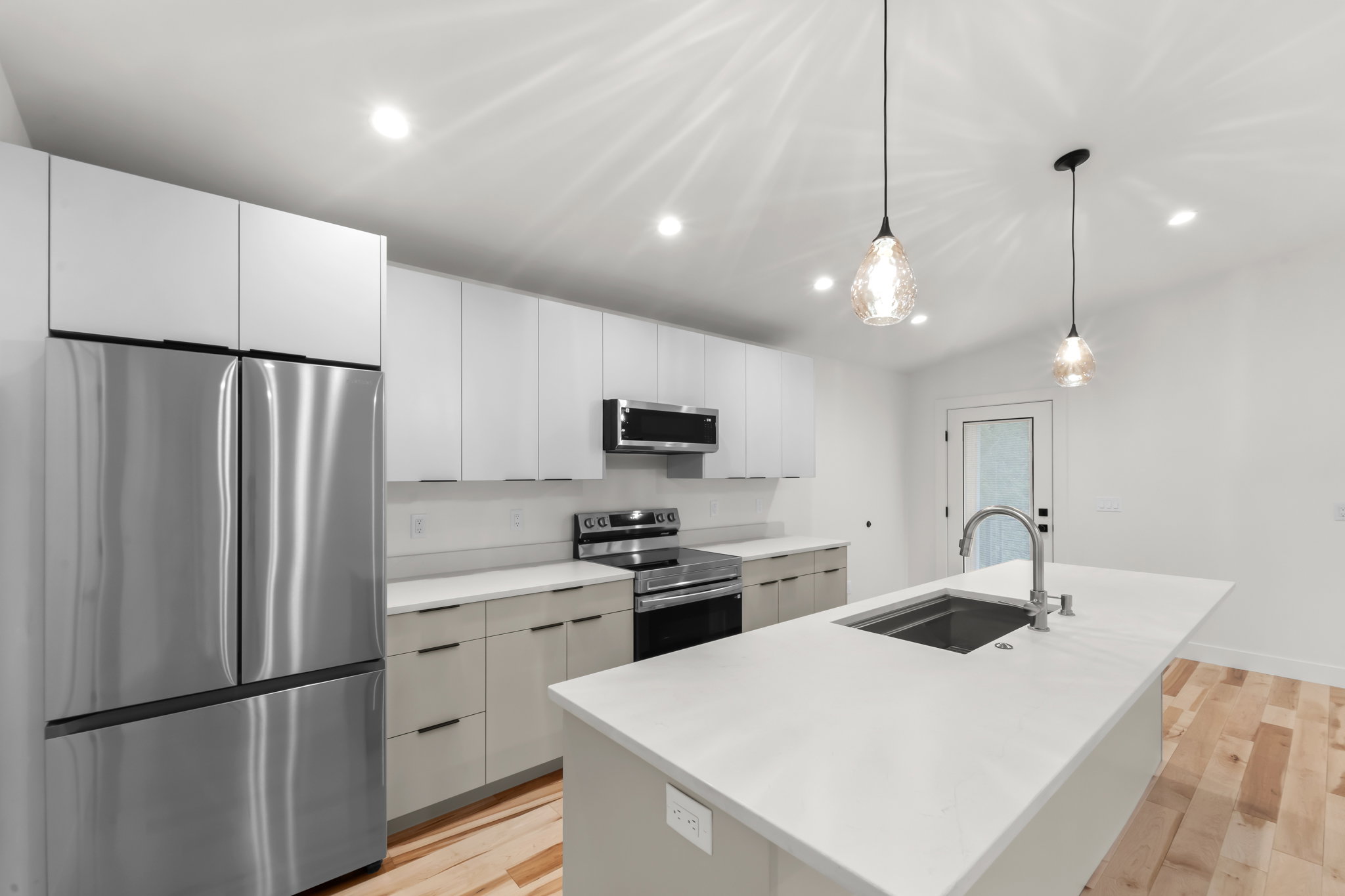 Modern kitchen featuring an expansive island with an undermount sink, stainless steel appliances, white flat-panel cabinetry, light wood-finish flooring, and recessed ceiling lighting