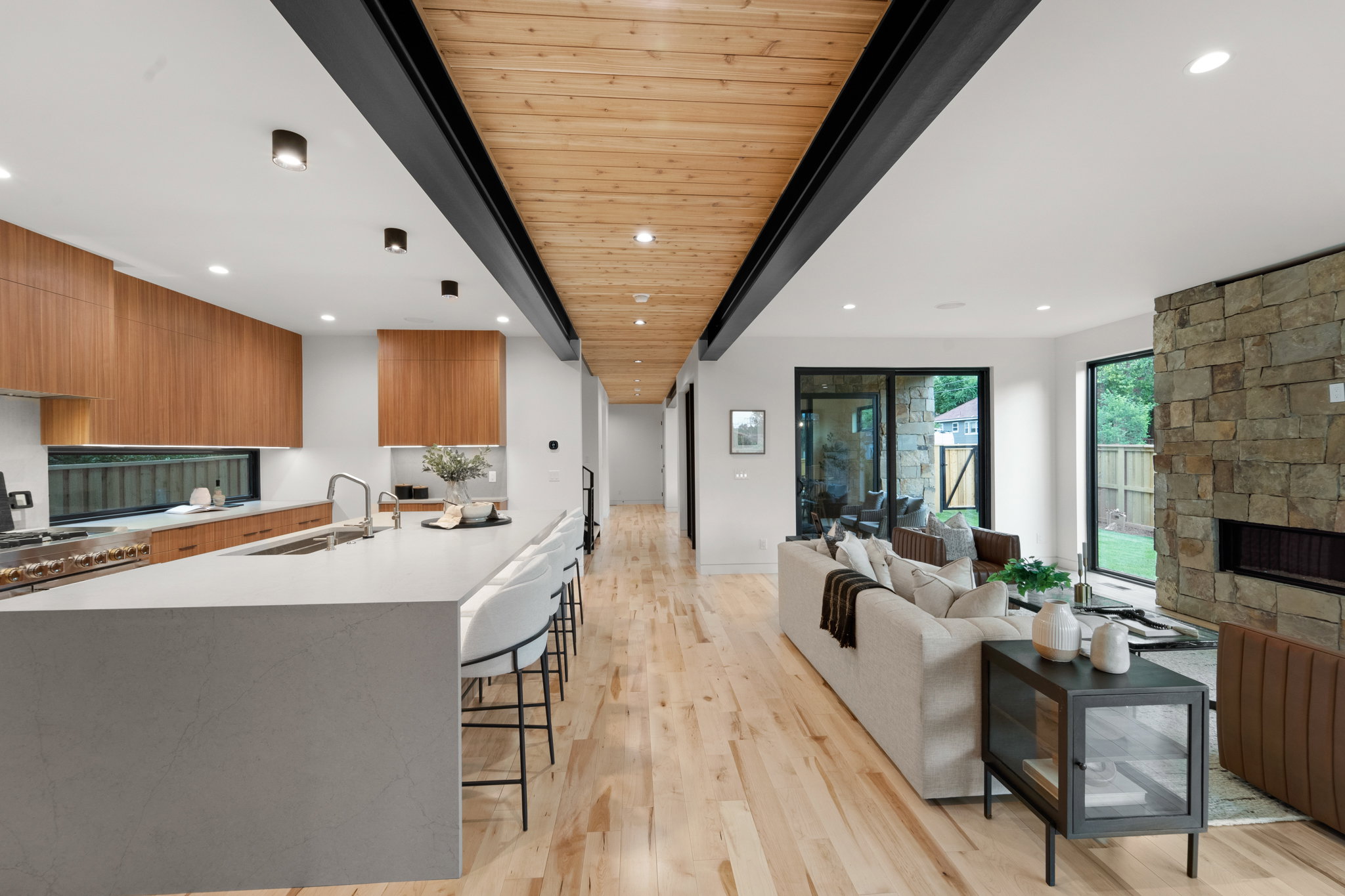 Open-concept living space featuring wood-finish flooring, a stone-clad fireplace, and a wood plank ceiling with exposed black beams