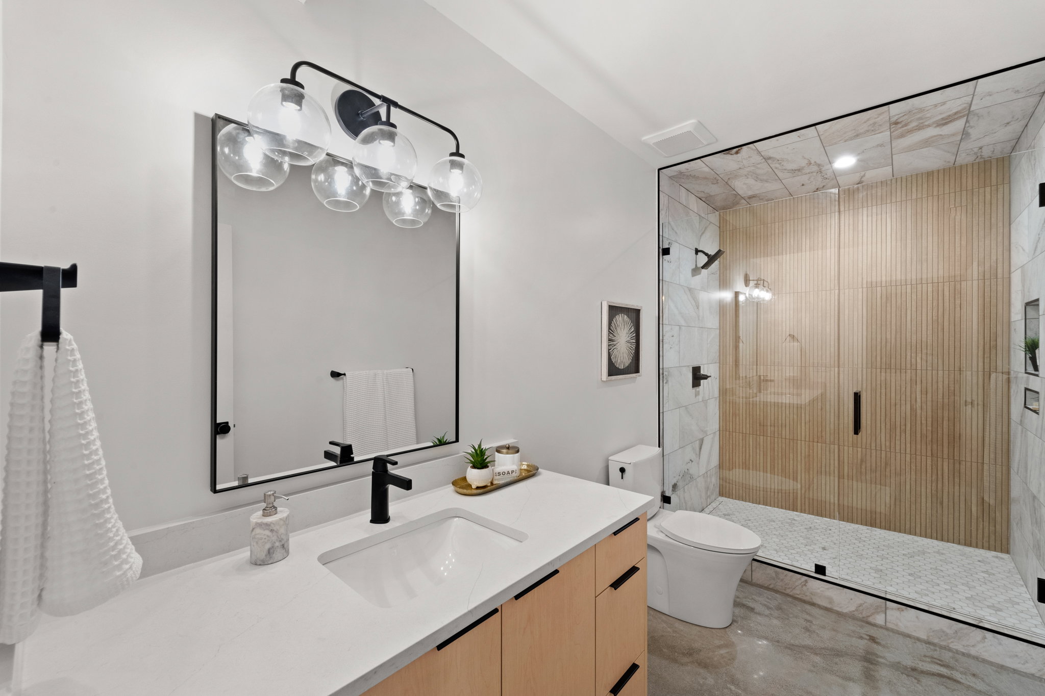 Bathroom featuring a frameless glass shower enclosure, wood-finish vanity with flat panel doors, matte black hardware, and a white countertop with integrated sink