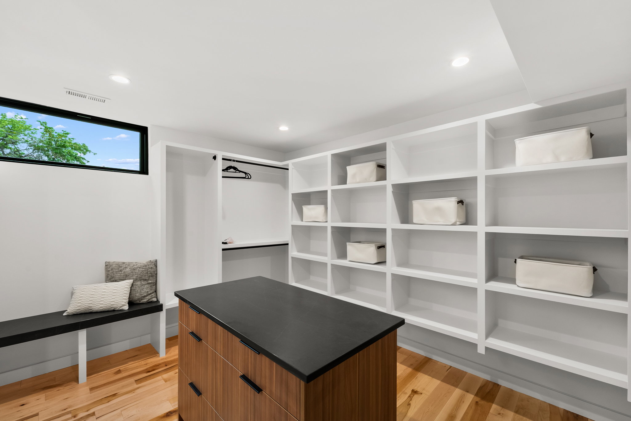 Custom built-in shelving, wood-finish flooring, a built-in bench, a black-framed window, and a central wood-tone island with a dark countertop