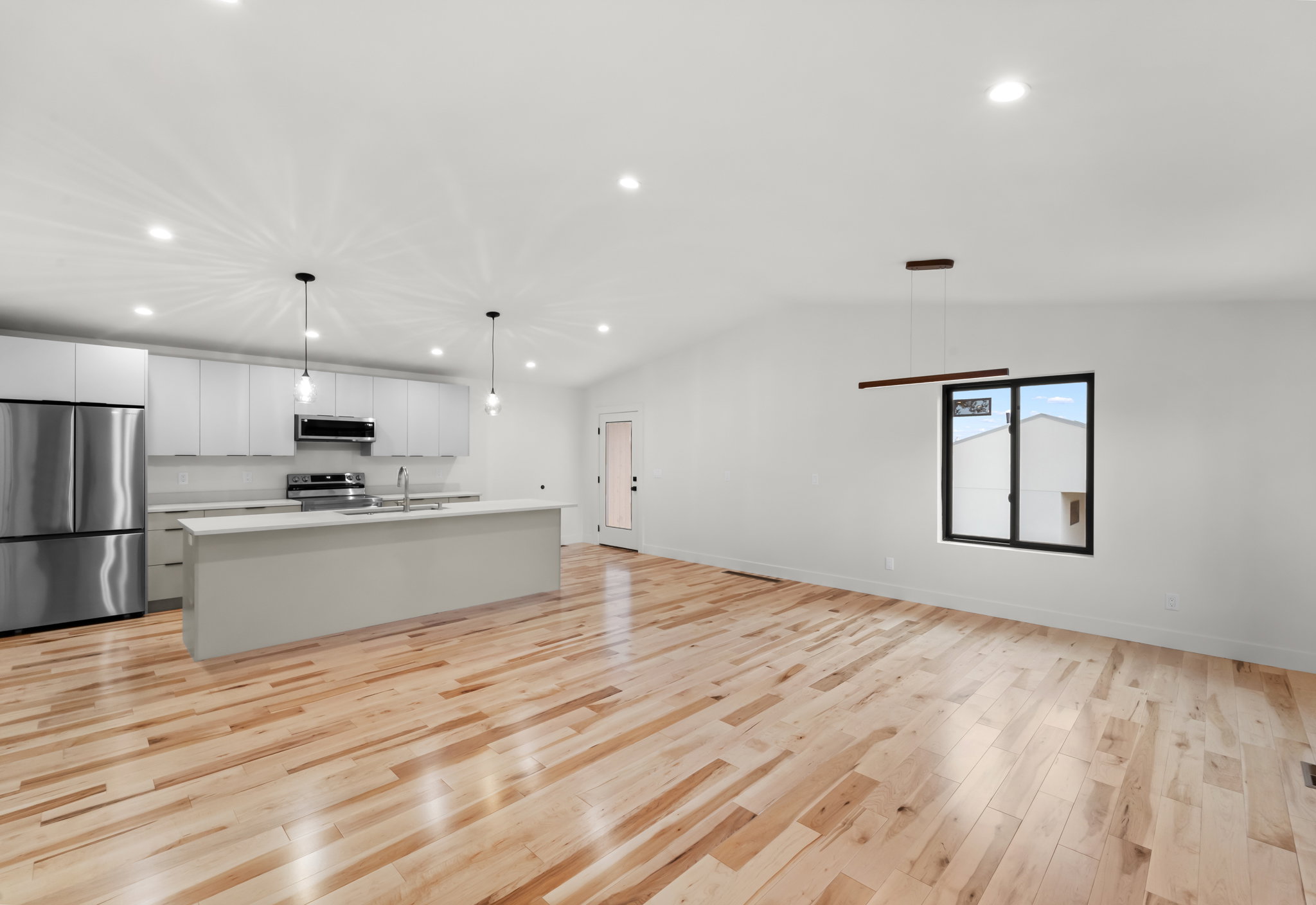 Open-concept living space featuring wood-finish flooring, a contemporary kitchen island with pendant lighting, stainless steel appliances, and a vaulted ceiling with recessed lighting