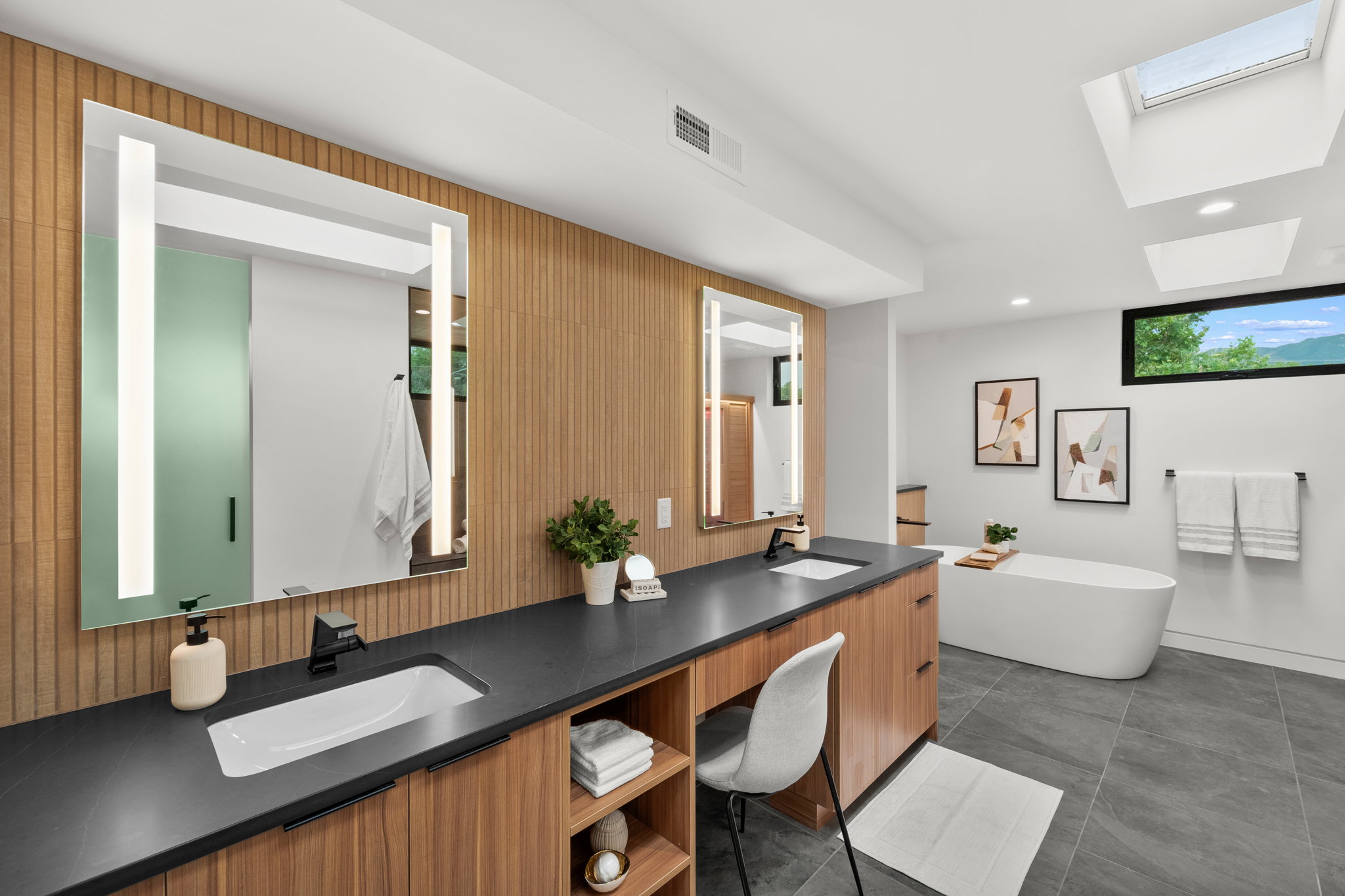 Spacious bathroom featuring a dual vanity with integrated lighting, wood-finish cabinetry, a freestanding soaking tub, and a skylight