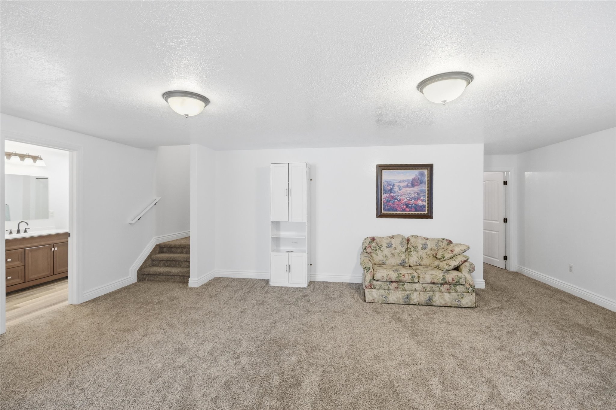 Spacious carpeted room featuring a white built-in cabinet with glass-front display, two flush-mount ceiling lights, and access to a bathroom with a wood-finish vanity and mirror