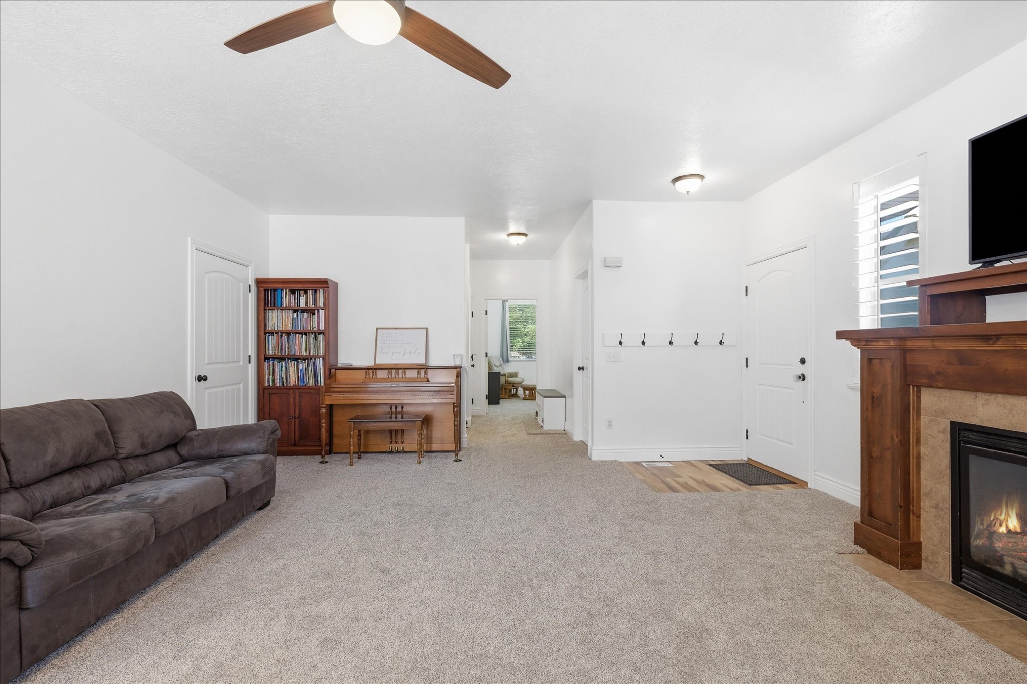 Carpeted living area featuring a fireplace with a wood-finish mantel and stone surround, a ceiling fan, and a front entry door with a coat rack
