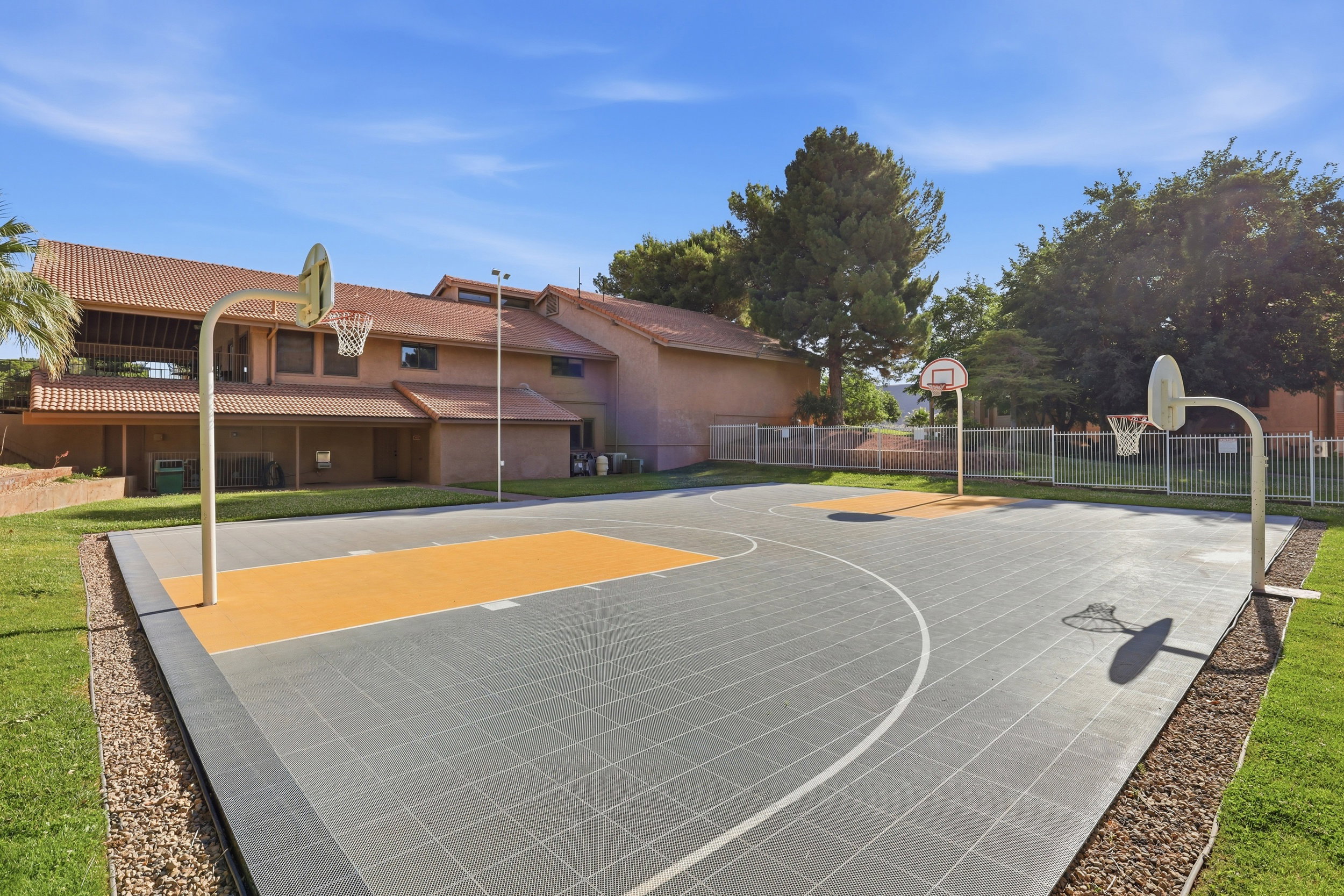 Outdoor basketball court featuring two hoops, a marked playing surface, and surrounding green space with gravel borders