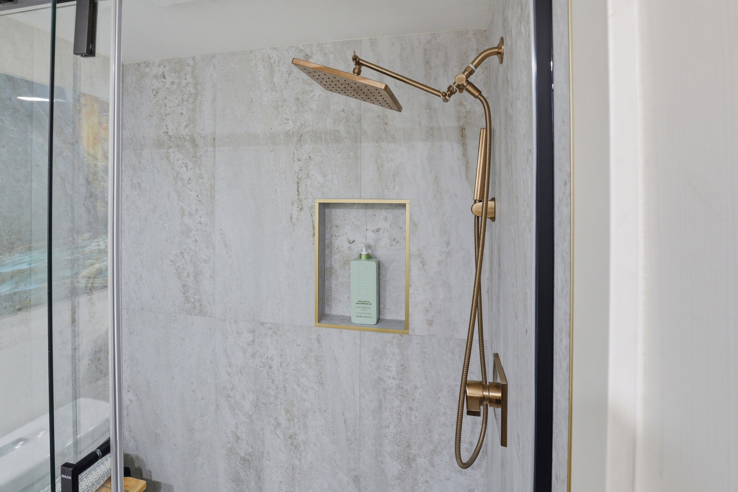 Spacious shower with large format tile, brushed bronze rain shower head, hand-held shower, and recessed shelf with gold-tone trim