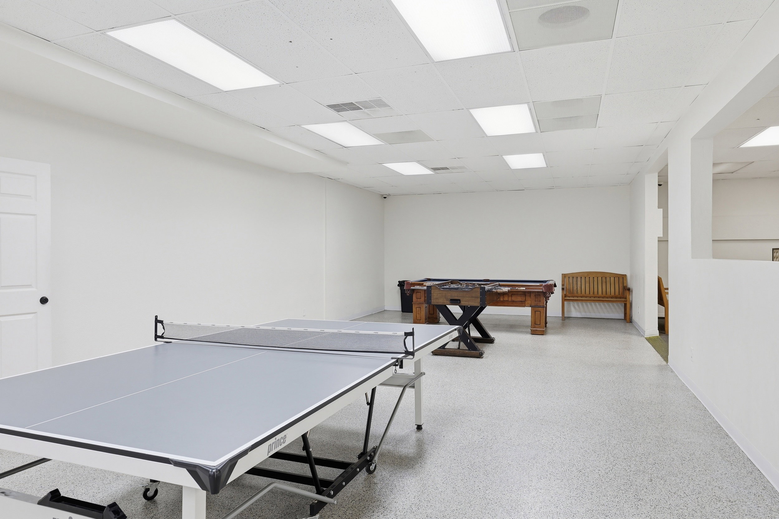 Spacious recreation area featuring white walls, a suspended ceiling with recessed lighting, and durable speckled flooring