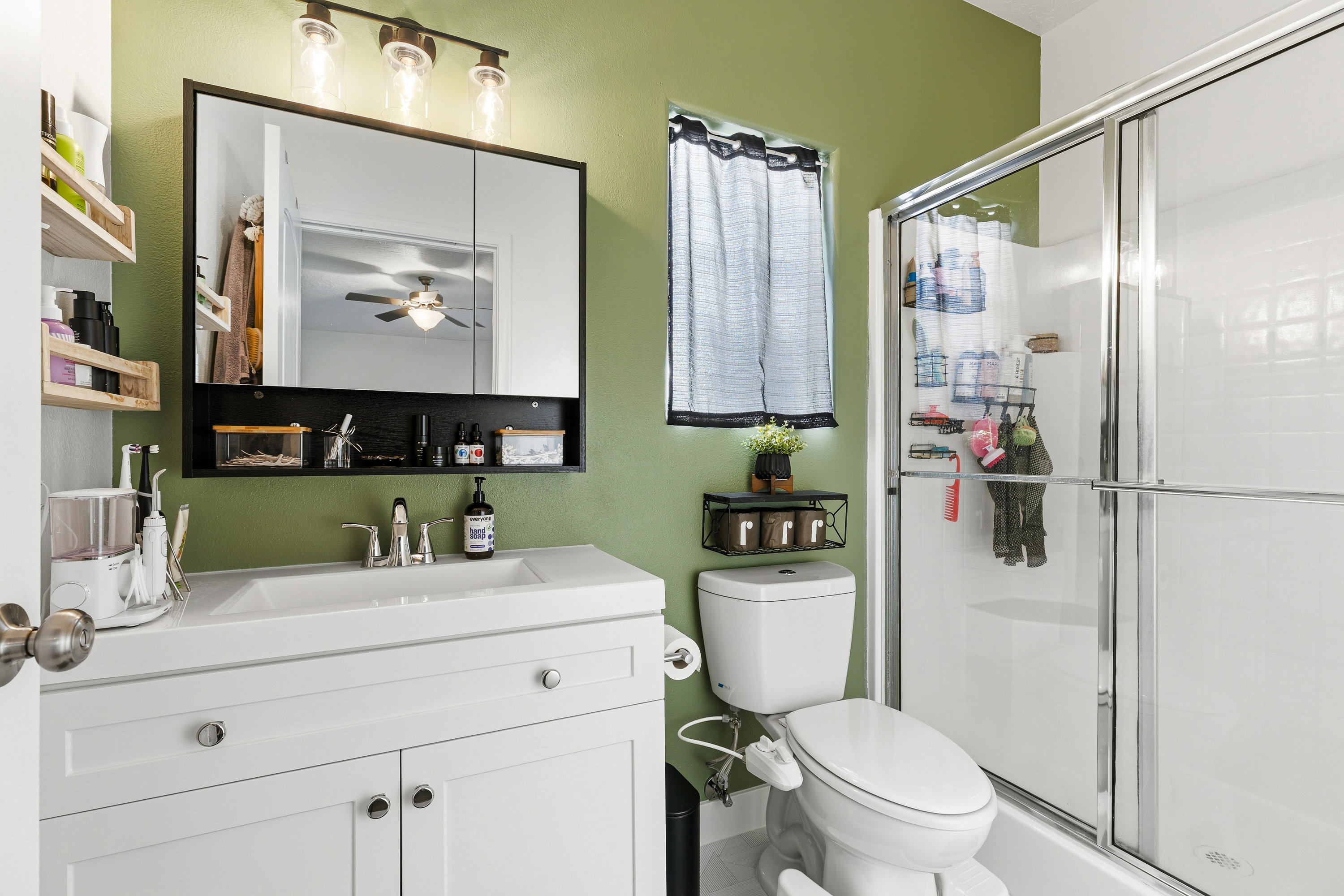 Bathroom featuring a single vanity with storage cabinetry, a framed mirror with integrated shelving, a three-light fixture, and a glass-enclosed shower