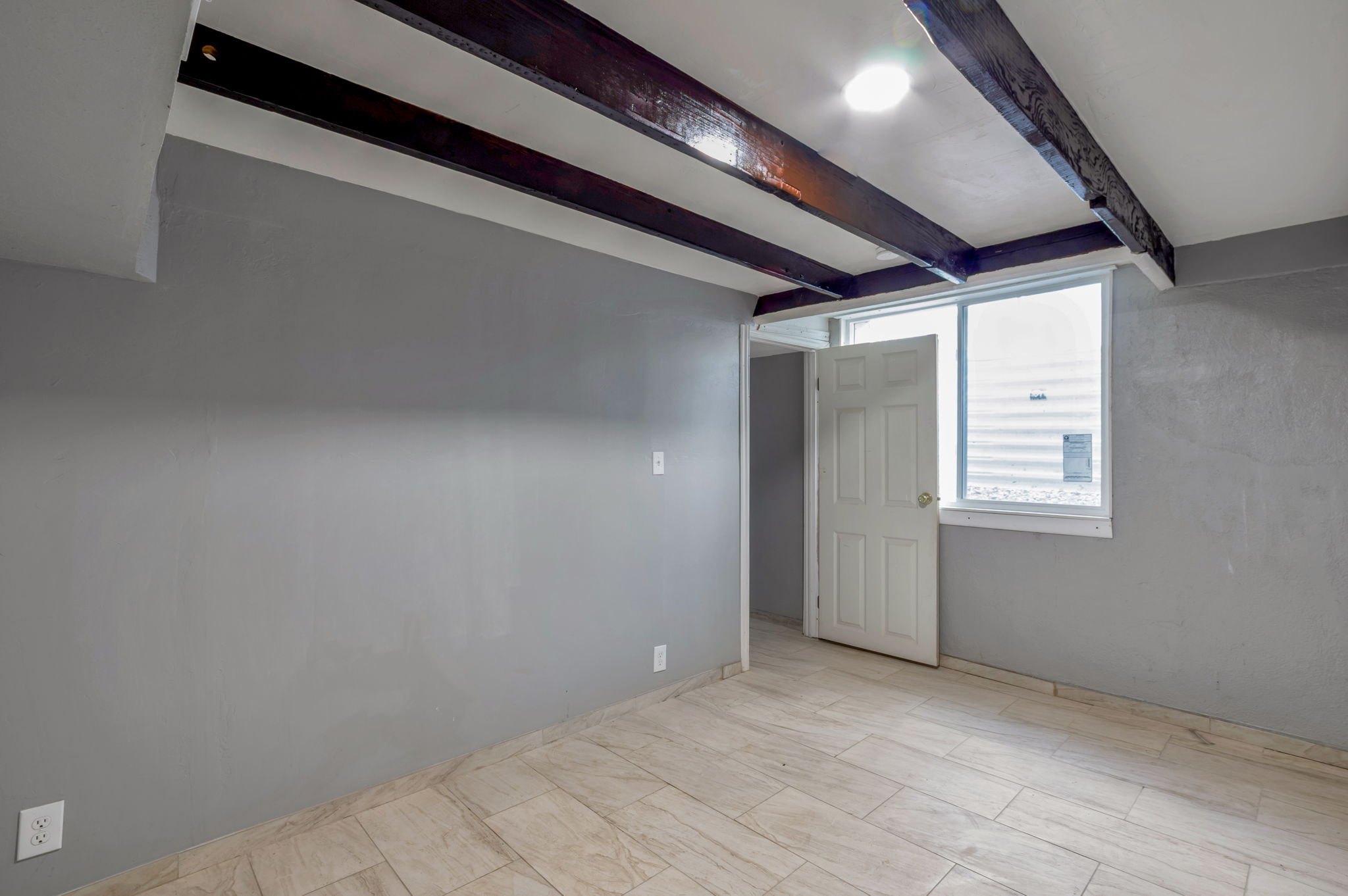 Room featuring exposed wood ceiling beams, light-colored tile flooring, gray painted walls, a white paneled door, and a single window