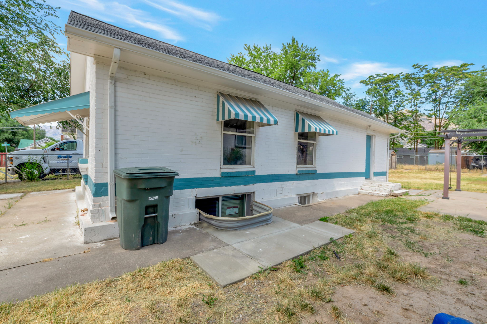 White brick exterior with teal trim, featuring two striped awnings over windows, a covered side entrance, and a basement window well