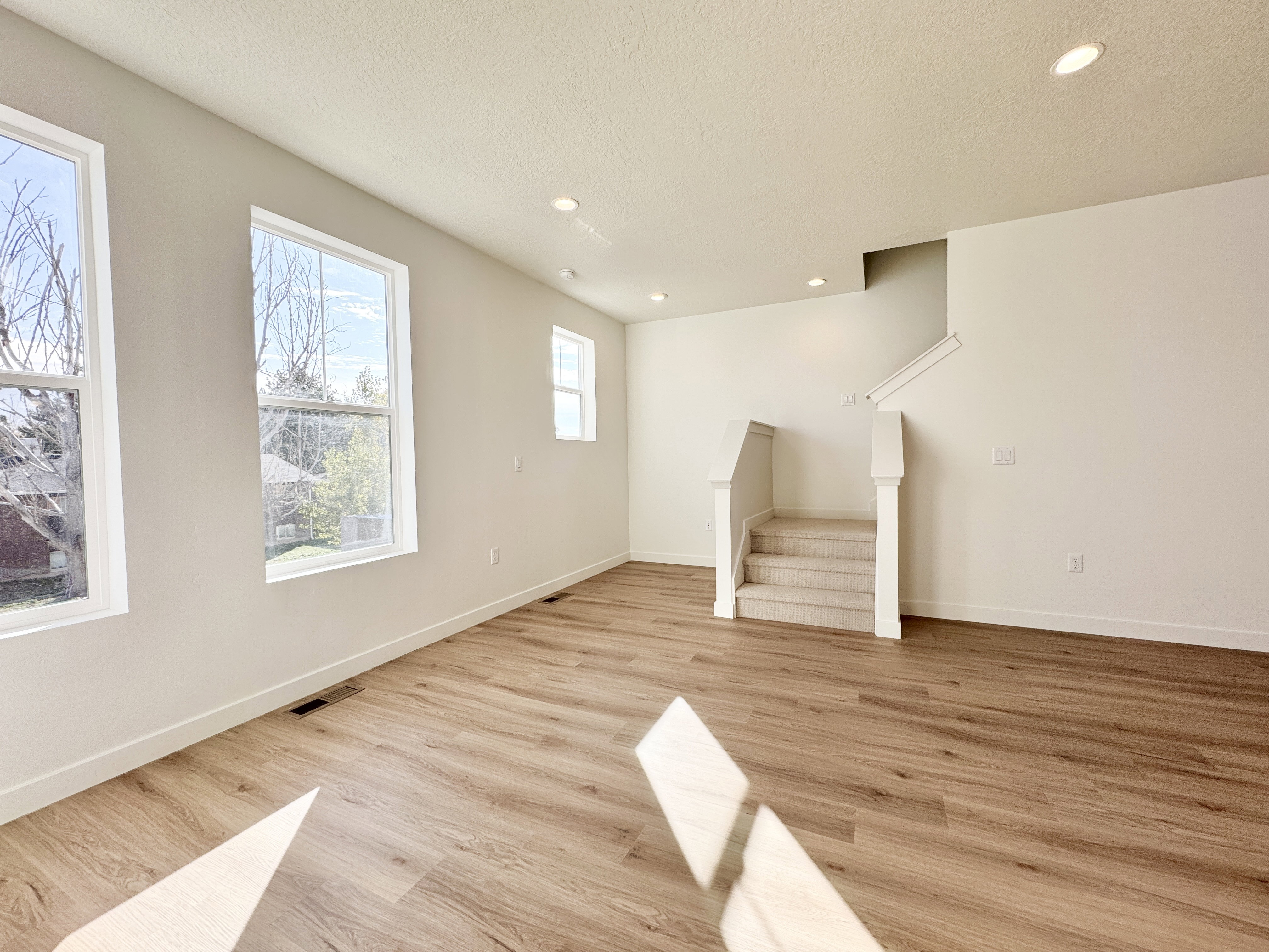 Spacious living area featuring large windows, light wood-finish flooring, recessed lighting, a staircase with carpeted treads, and crisp white walls