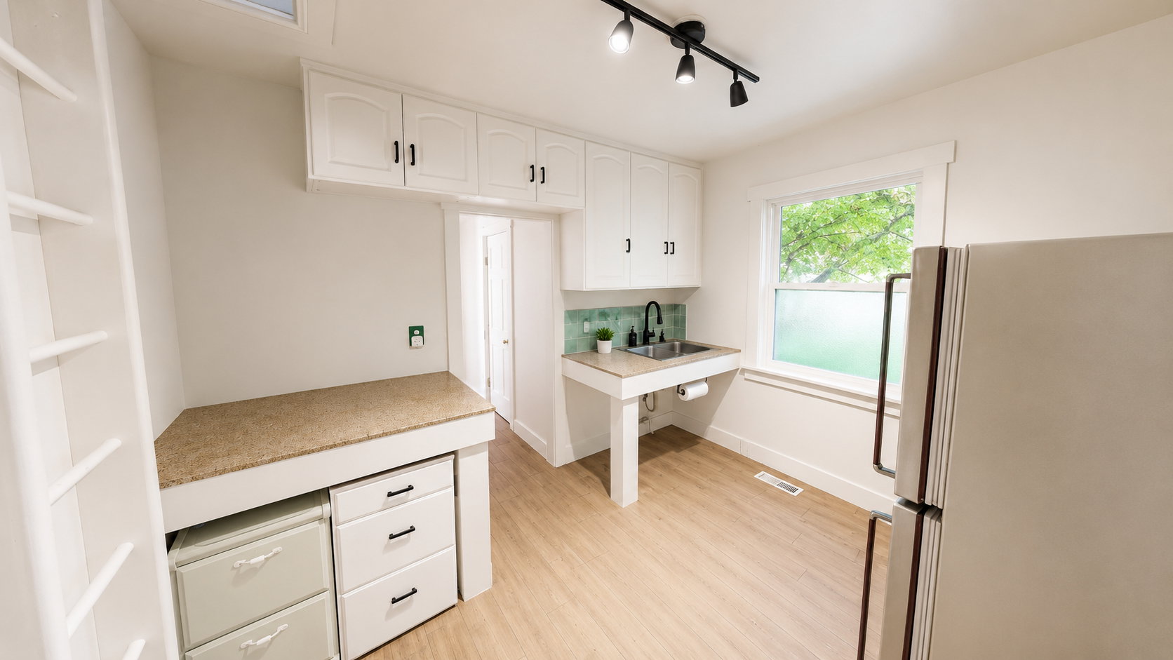 Compact kitchen space featuring light wood-finish flooring, white cabinetry with black hardware, a stainless steel sink with a matte black faucet, and a green tiled backsplash