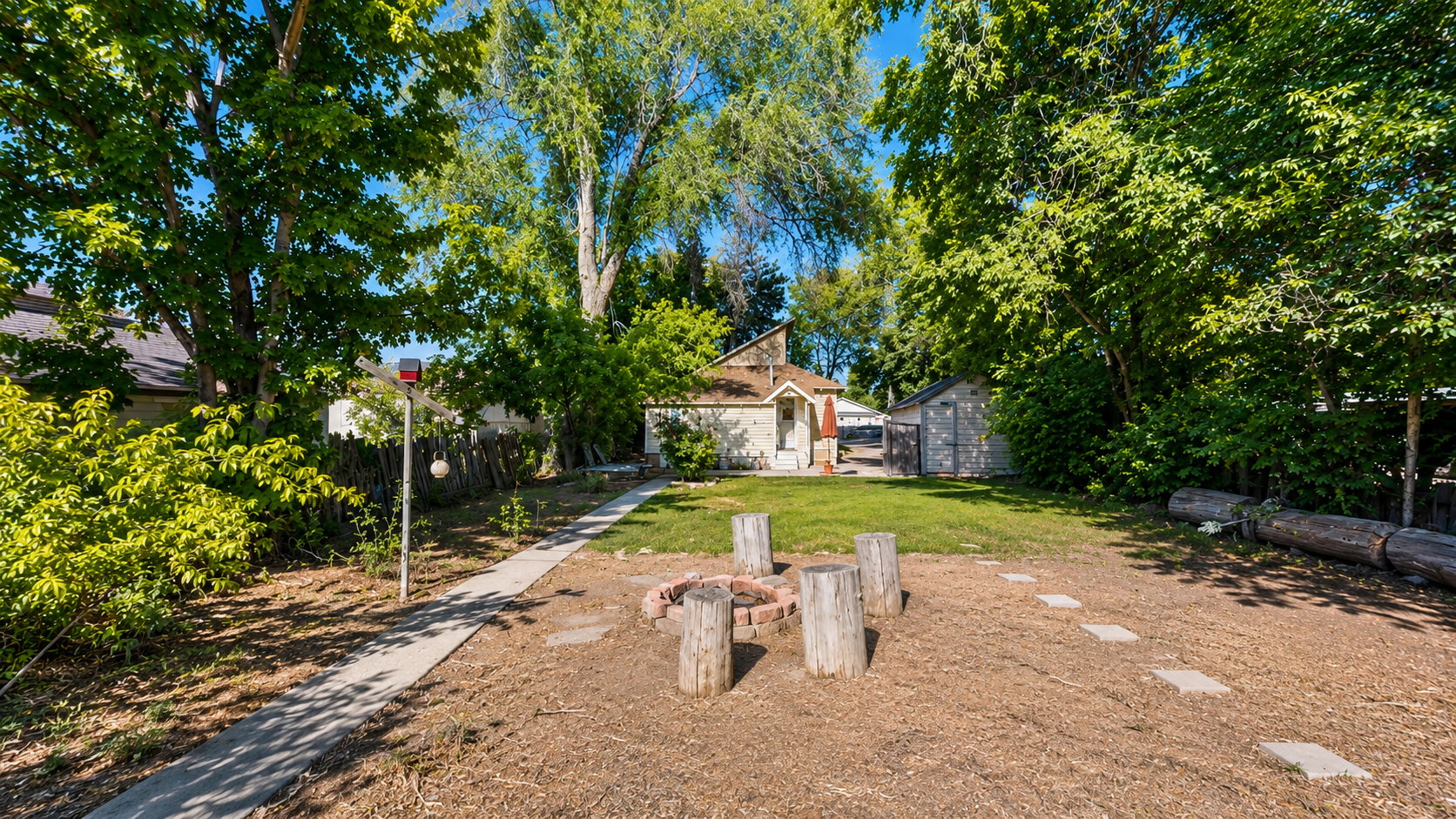 Expansive backyard featuring a stone fire pit with log seating, a manicured lawn area, and mature trees providing natural shading