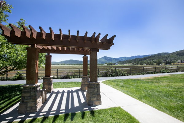 Wood-finish pergola with stone bases, manicured lawn, paved walkway, split-rail fencing, and distant mountain scenery