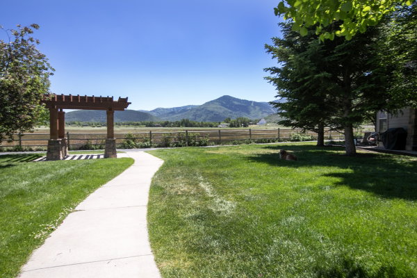 Expansive green lawn with a concrete pathway leading to a wood pergola with stone bases, featuring a wooden fence in the background and distant mountains
