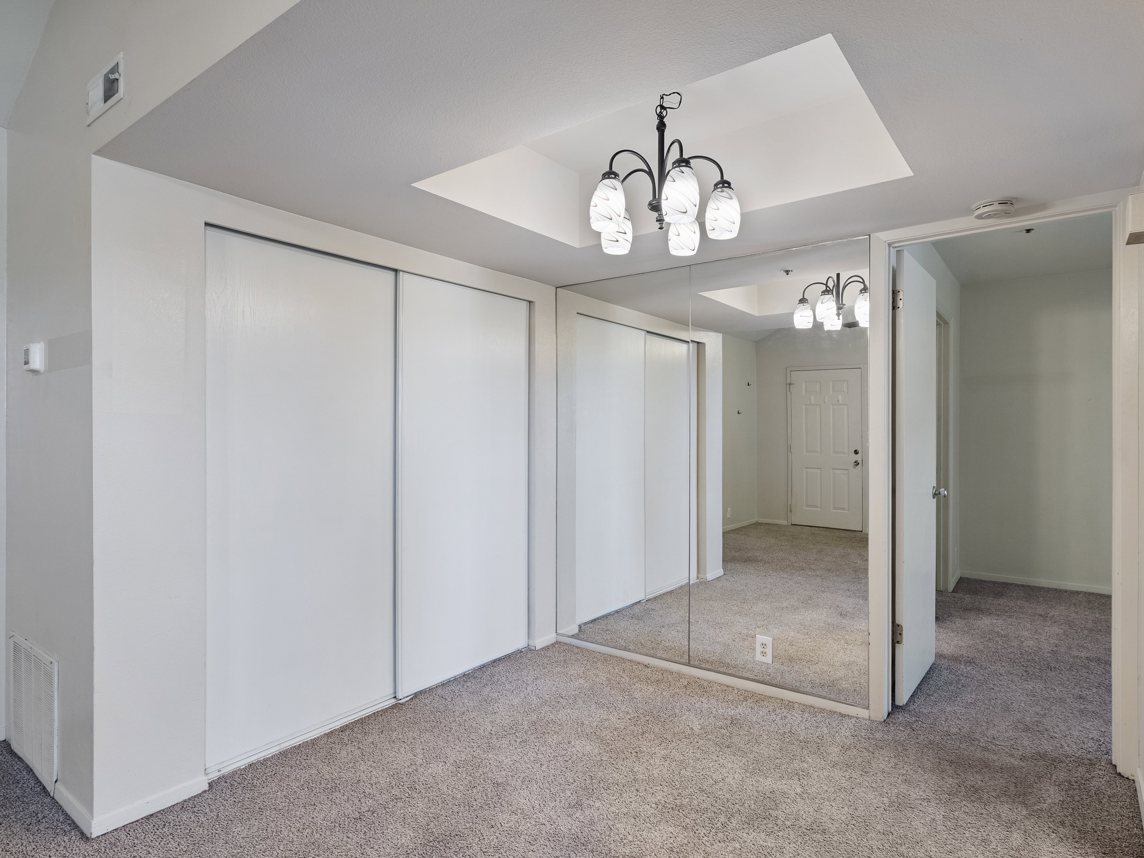 Recessed ceiling with an ornate chandelier, mirrored wall panels, white sliding closet doors, and light gray carpeting