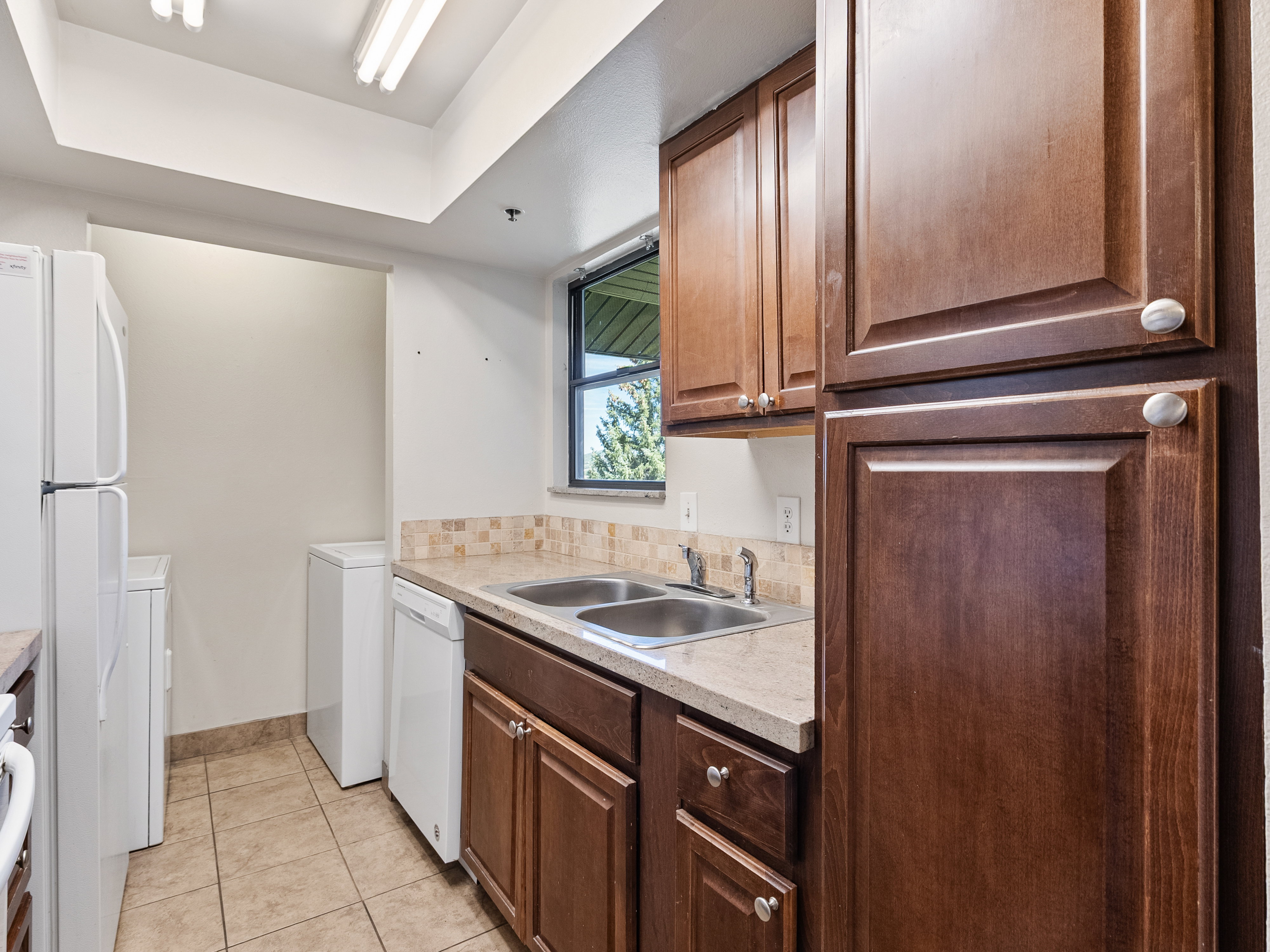 Kitchen area featuring wood-finish cabinetry, a double basin stainless steel sink, light-toned countertop, and a tiled backsplash