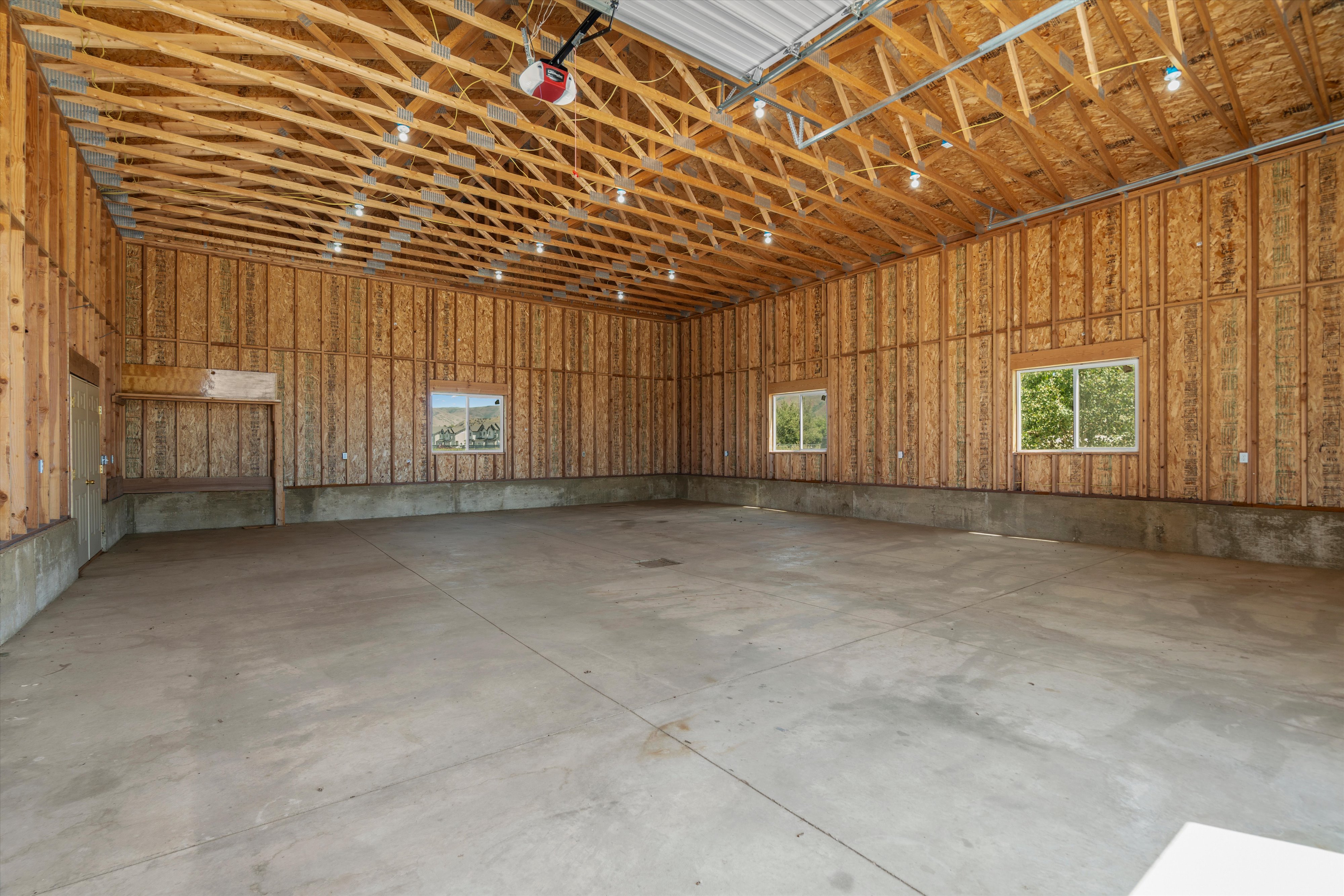 Spacious garage with exposed wood framing, concrete flooring, multiple windows, overhead lighting, and an automatic garage door opener