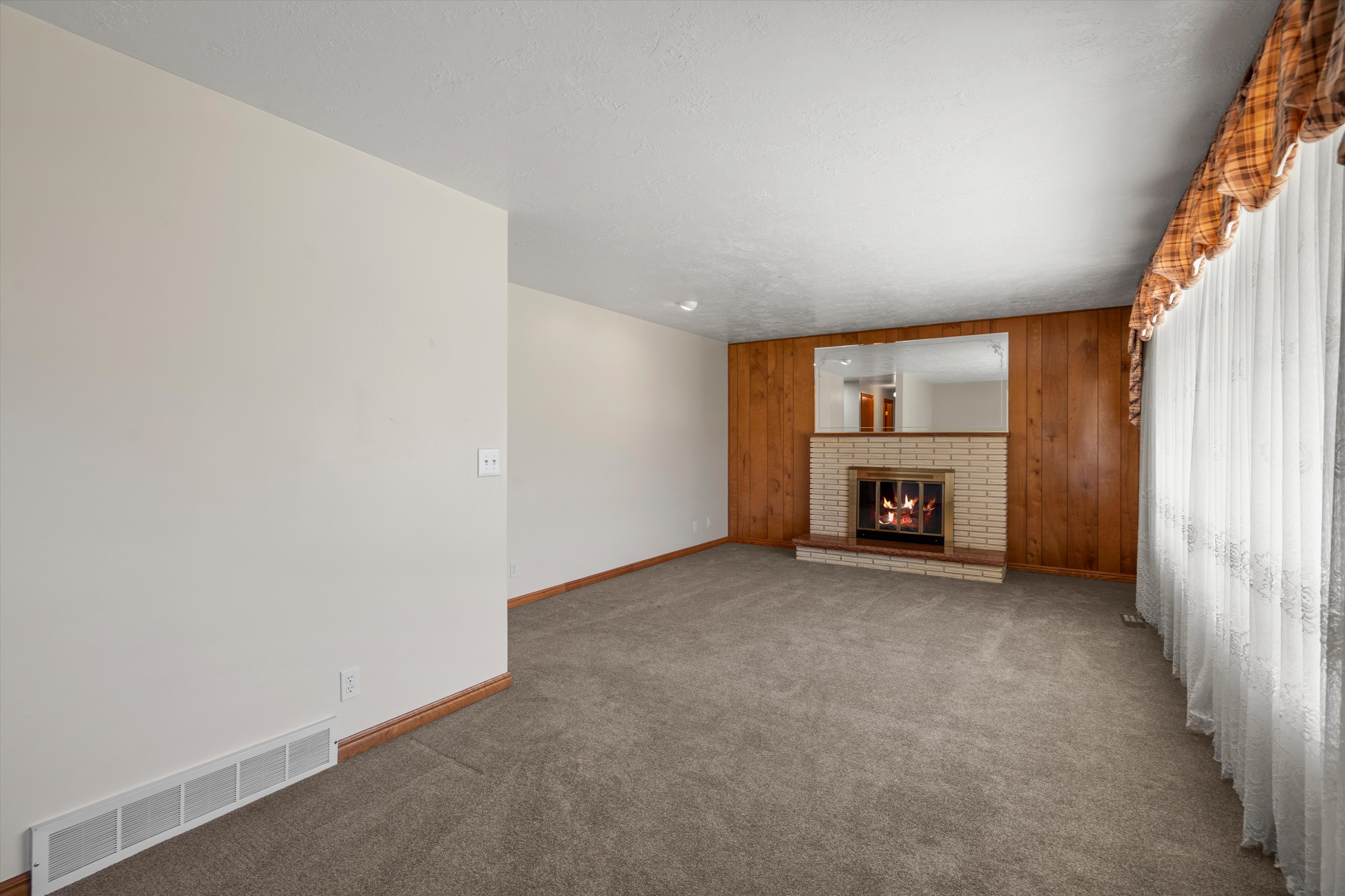 Carpeted living area featuring a brick fireplace with a wood-paneled accent wall, a mirrored overmantel, and window treatments with sheer curtains