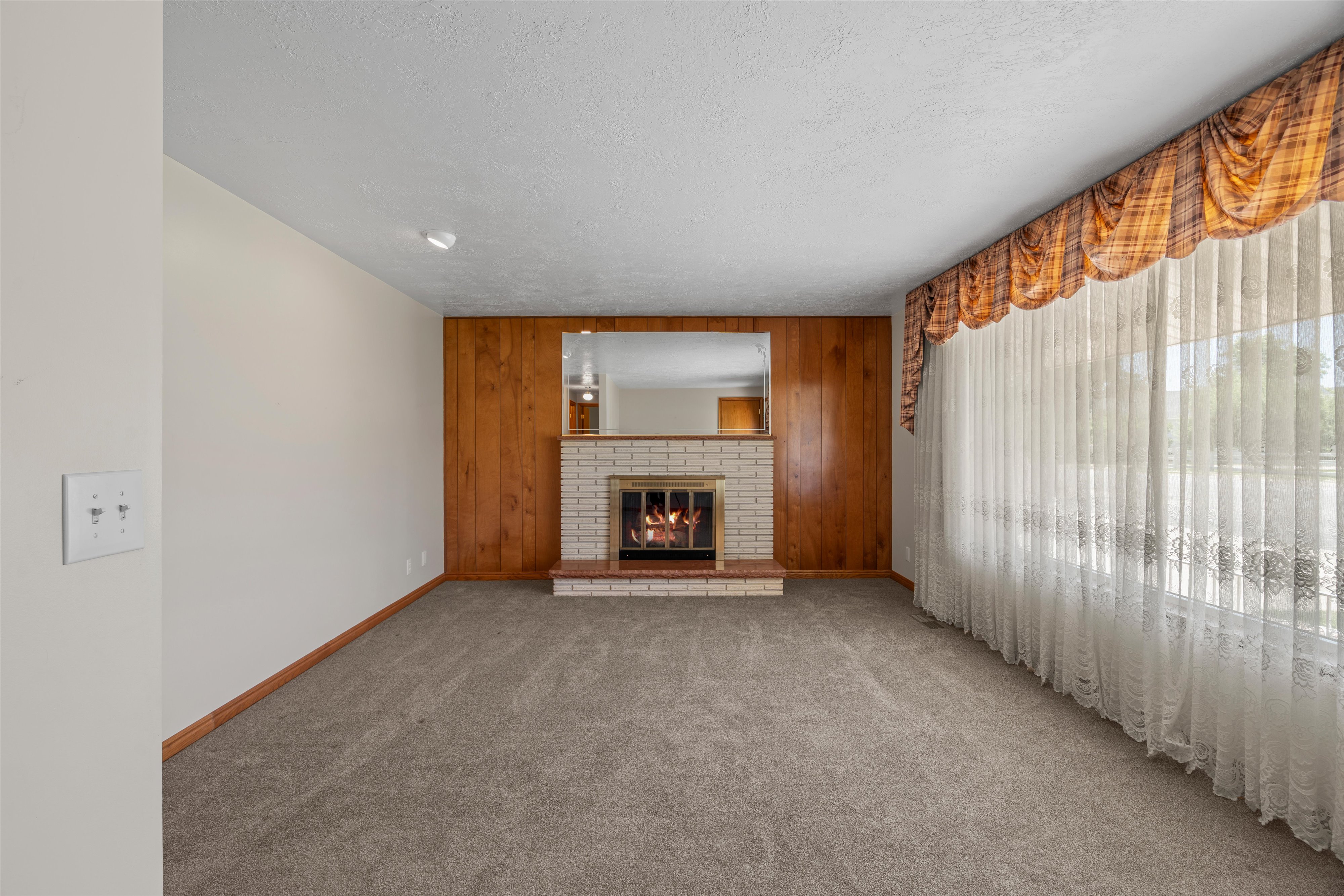 Living room featuring wood paneling, a brick fireplace, and a large window with sheer drapery