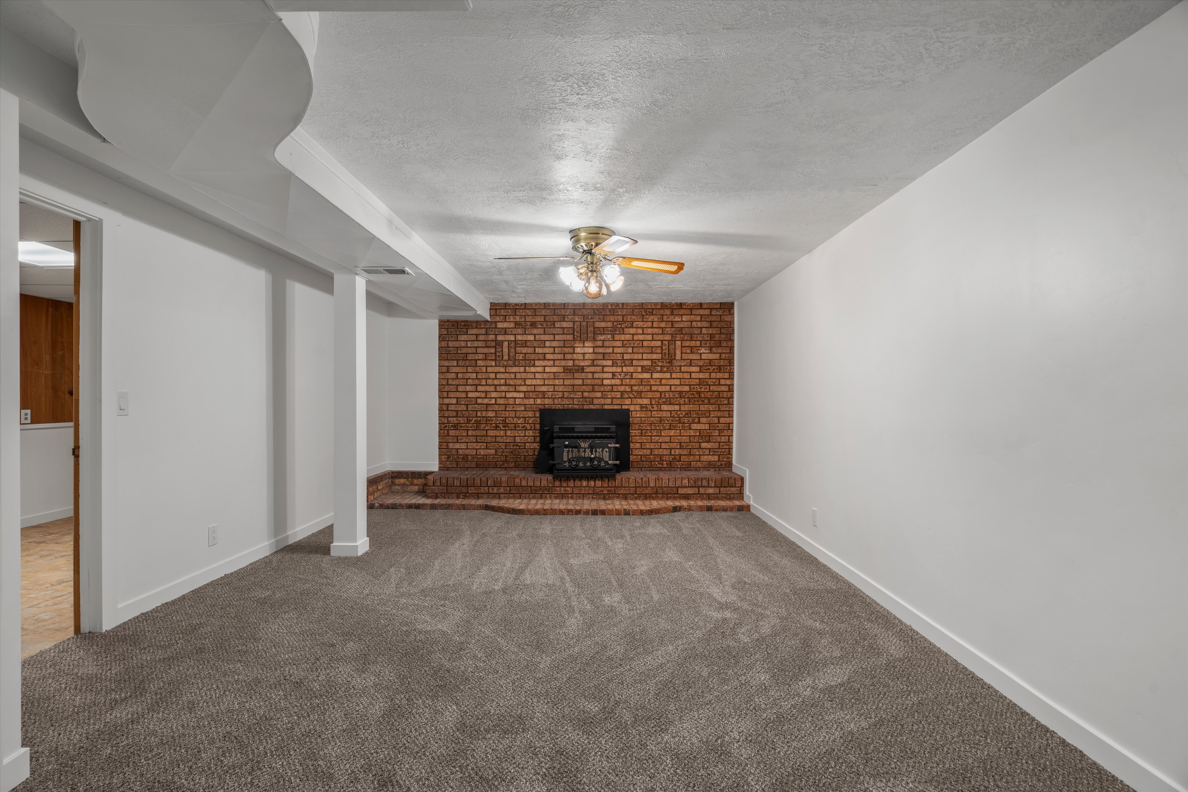 Spacious room featuring a brick fireplace with a raised hearth, gray patterned carpeting, a ceiling fan with integrated lighting, and white baseboards