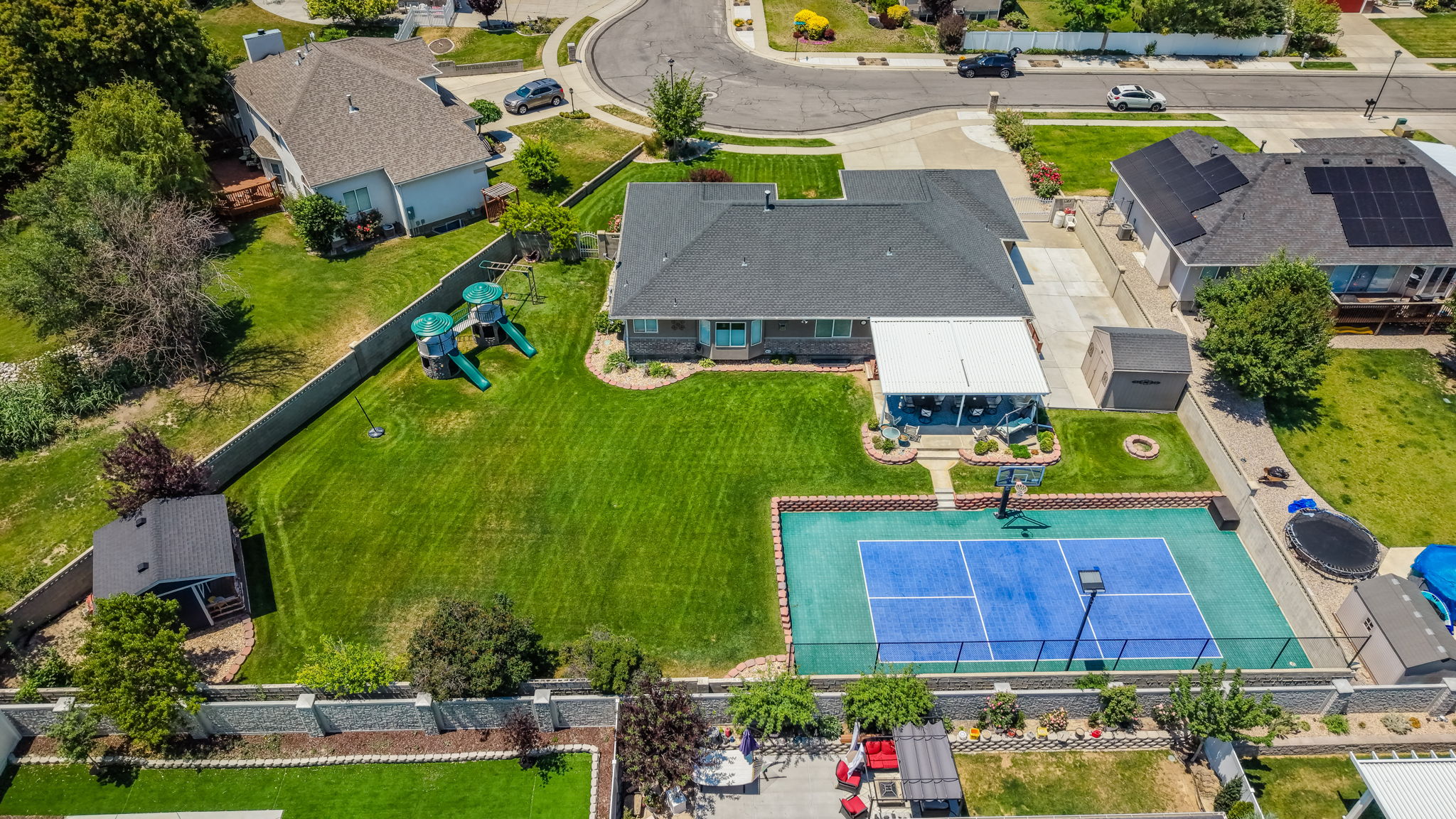 Expansive backyard featuring a blue multi-sport court, a covered patio, a manicured lawn, and mature trees