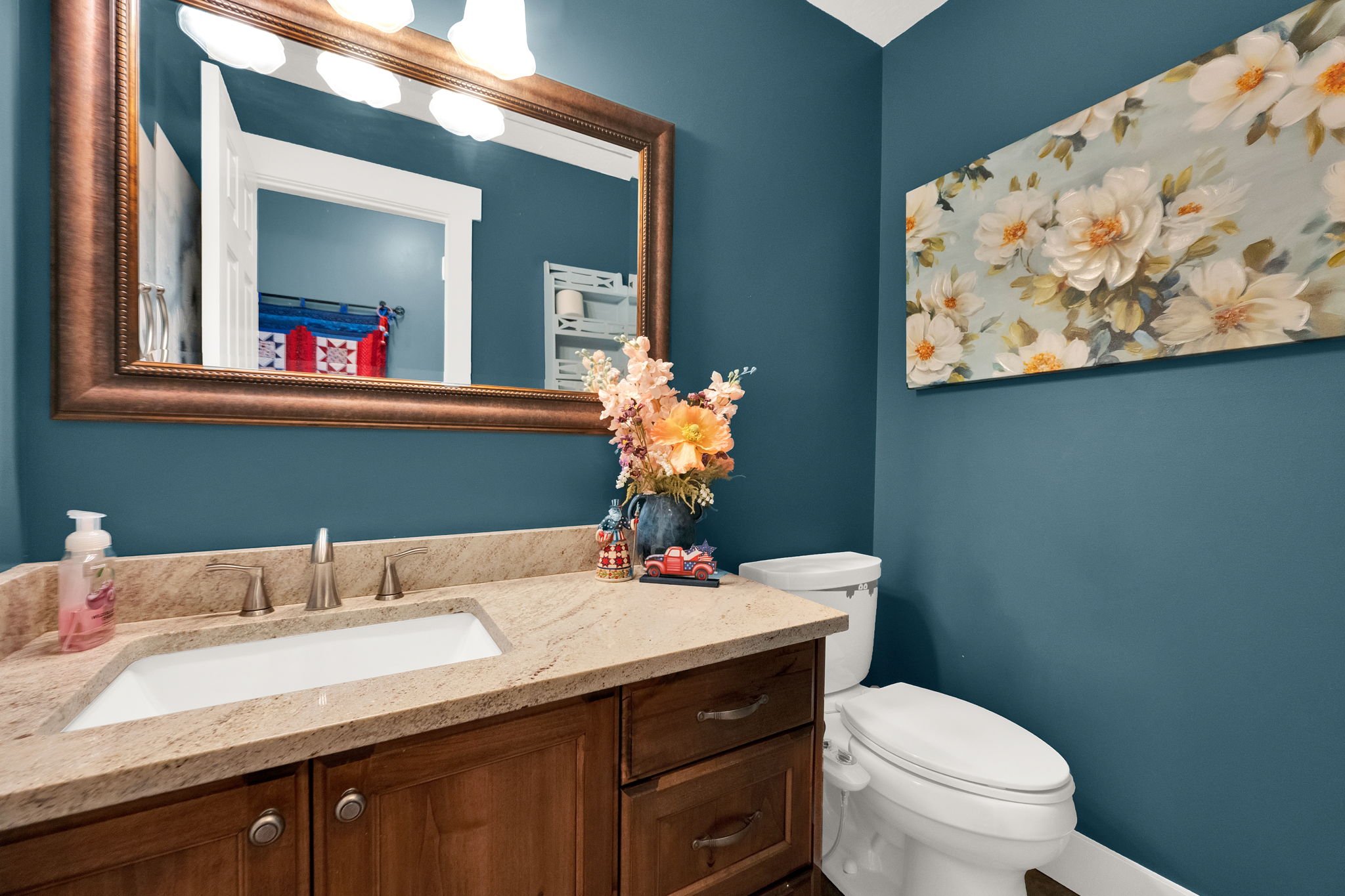 Bathroom vanity featuring a light-toned stone countertop with an undermount sink, a bronze-finish framed mirror with attached overhead lighting, and rich wood-finish cabinetry