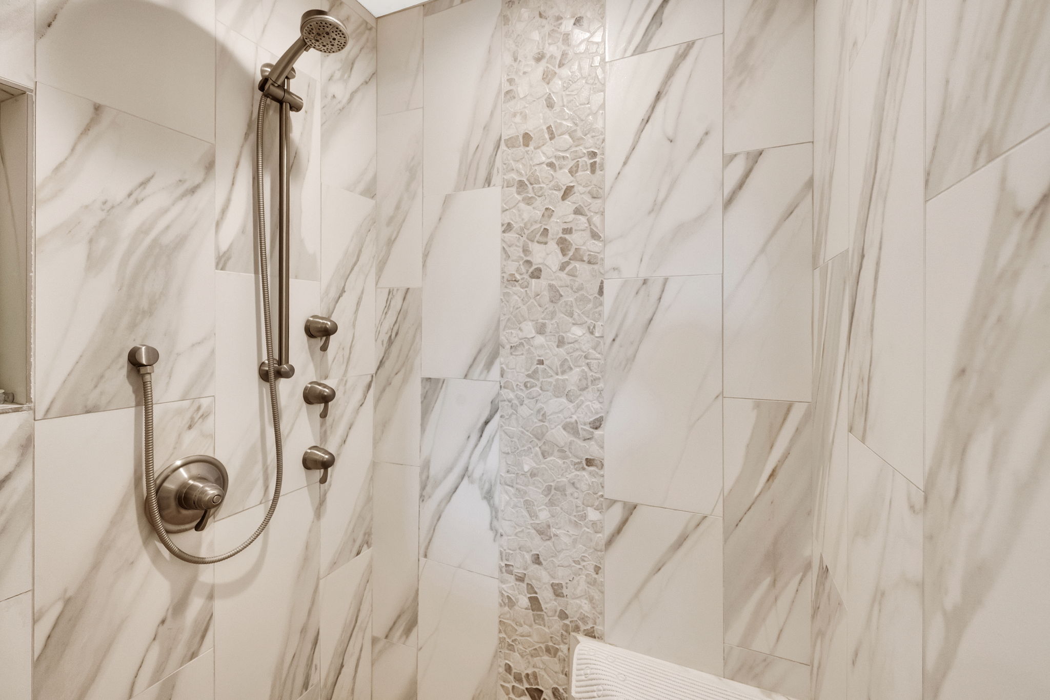 Shower stall featuring large format marble-look tile, a pebble tile accent strip, and brushed nickel fixtures including a rainfall shower head and body sprays