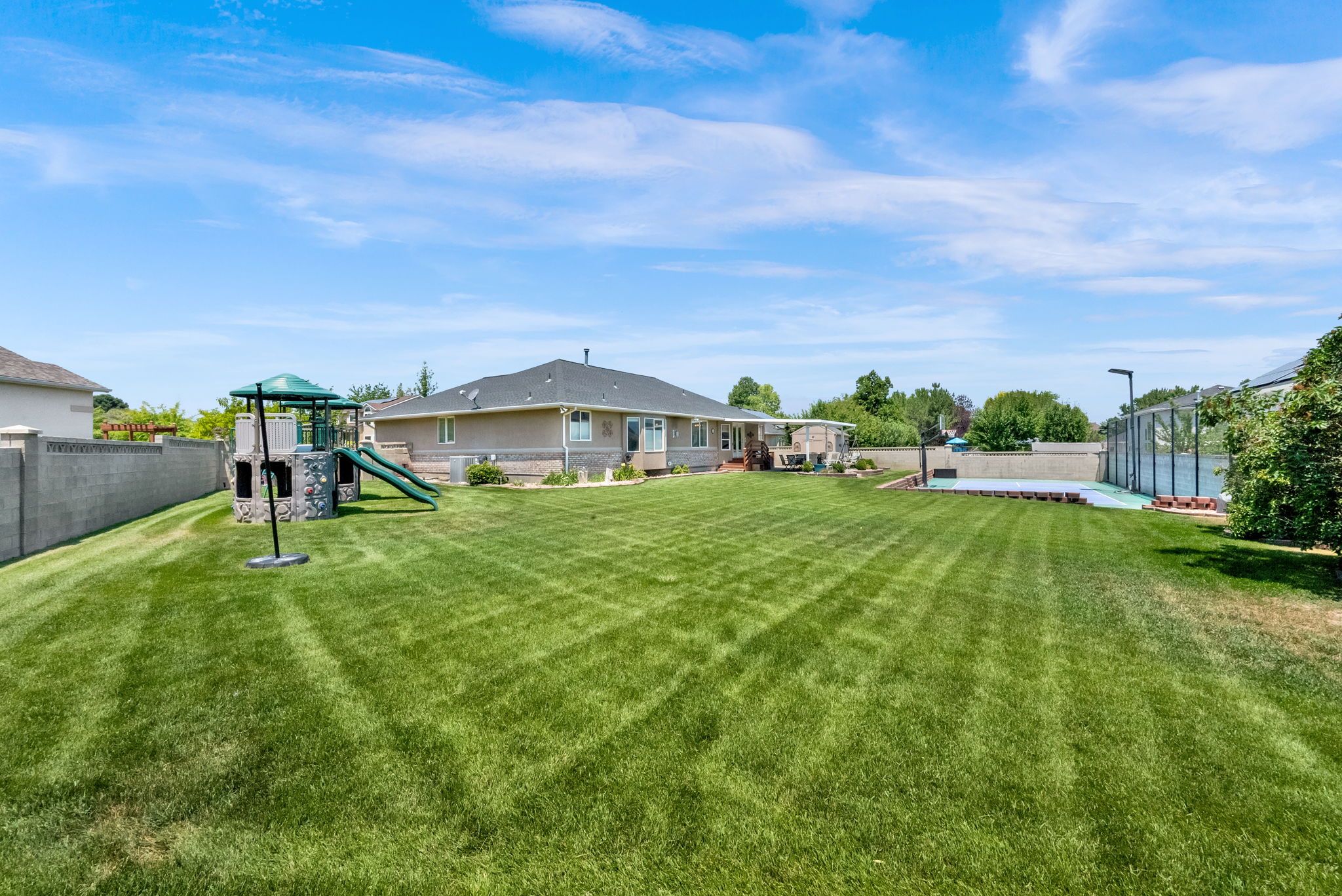 Expansive backyard featuring a lush lawn, a dedicated play structure with slides, a tennis court, and a perimeter fence