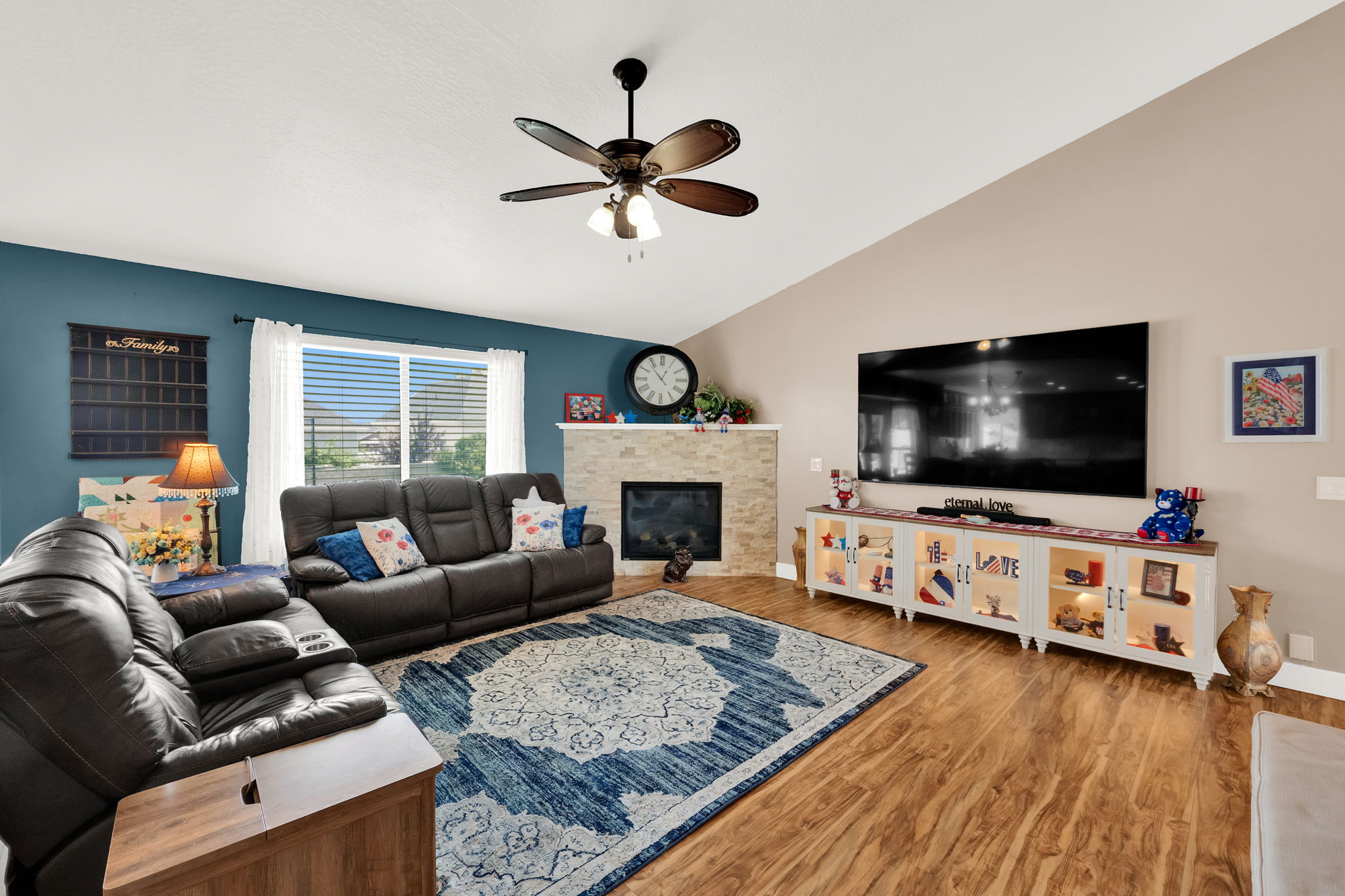 Vaulted ceiling living area featuring a stone-clad fireplace, wood-finish flooring, and a ceiling fan
