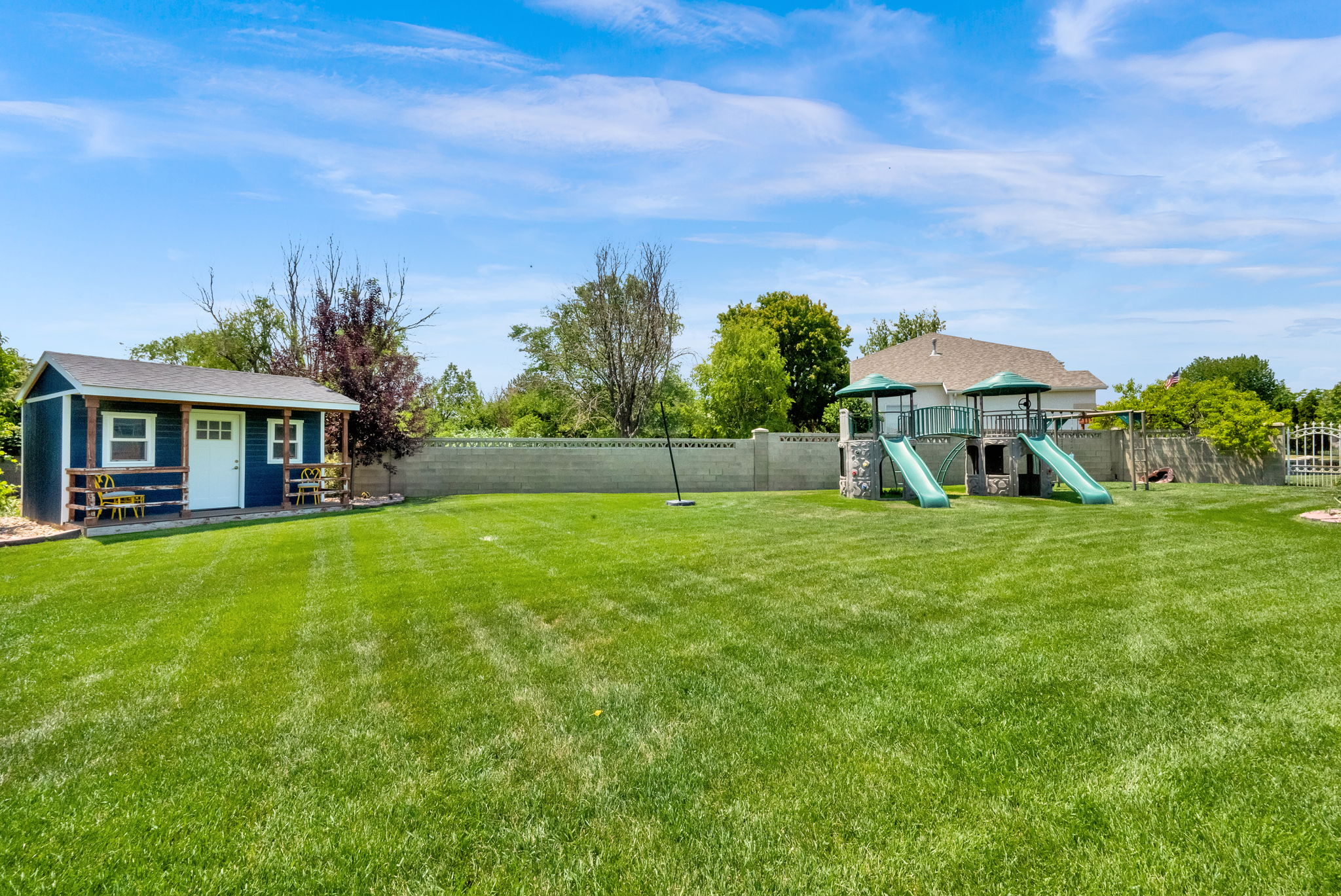 Expansive lawn area featuring a detached storage shed with a covered porch, a stone-accented play structure with dual slides, and a perimeter fence