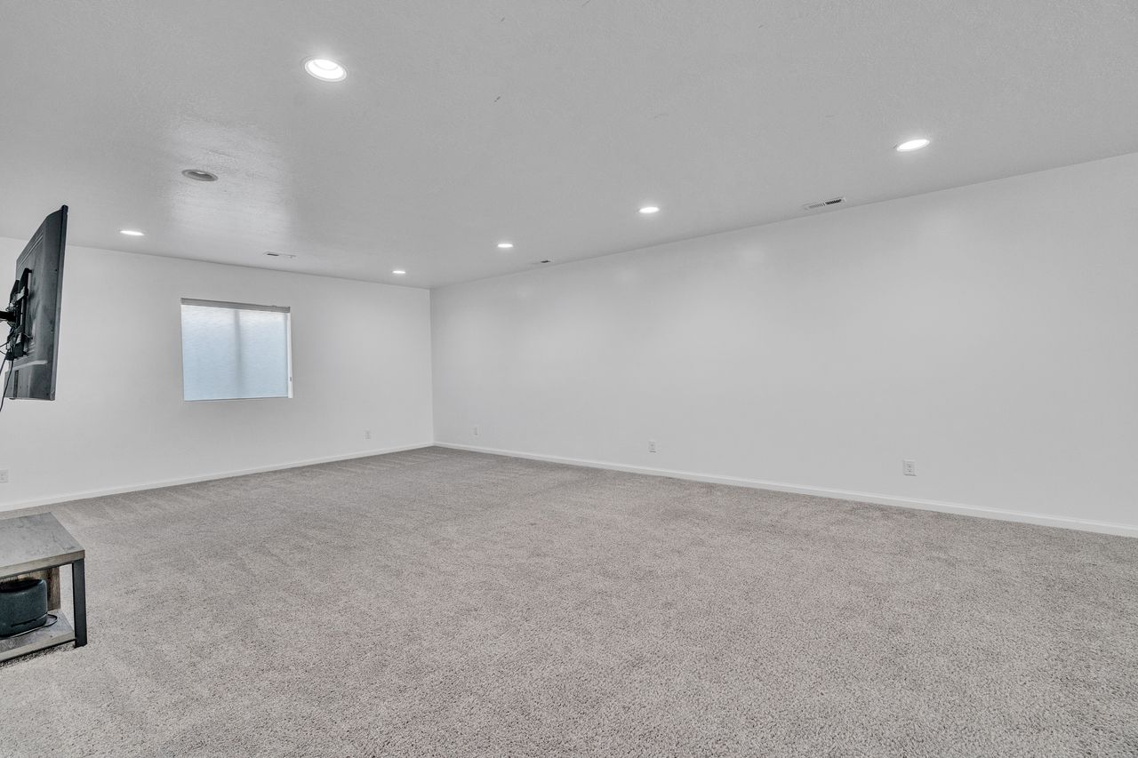 Spacious room featuring light gray carpeting, recessed ceiling lighting, a single window with a privacy screen, and white painted walls