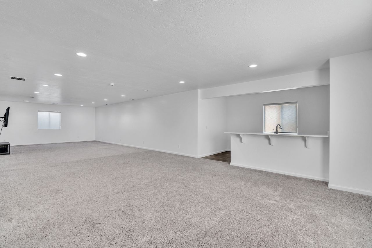 Spacious room featuring light gray carpeting, recessed ceiling lighting, a built-in bar with a sink and window, and white walls