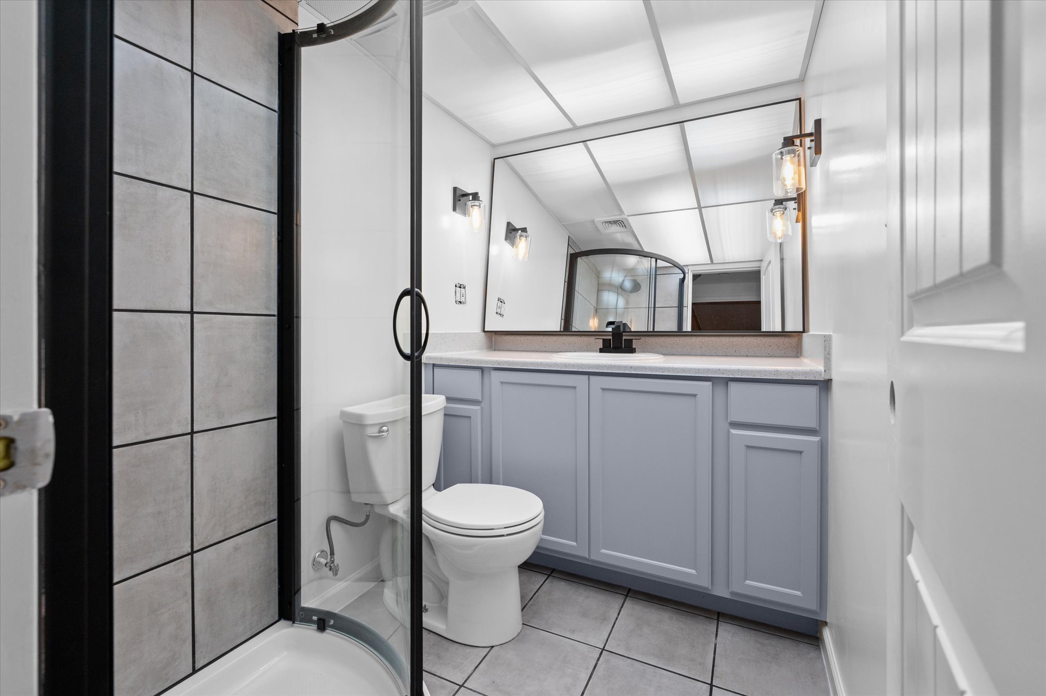Contemporary bathroom featuring a curved glass shower enclosure with dark framing, a single vanity with gray cabinetry and light countertop, and a large wall-mounted mirror
