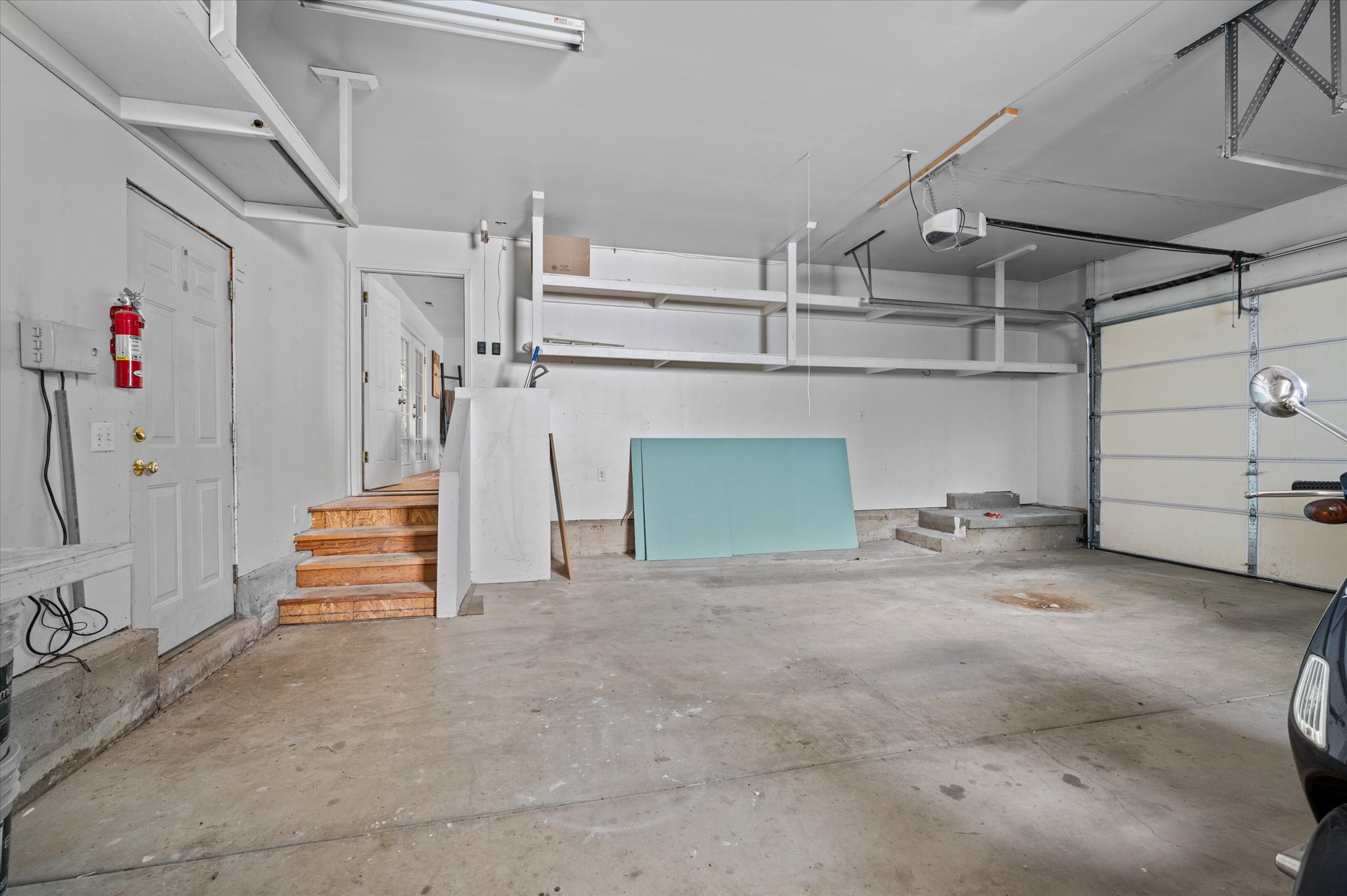 Spacious garage featuring concrete flooring, built-in overhead storage shelves, a full-panel garage door, and interior access via a paneled door with wooden stairs