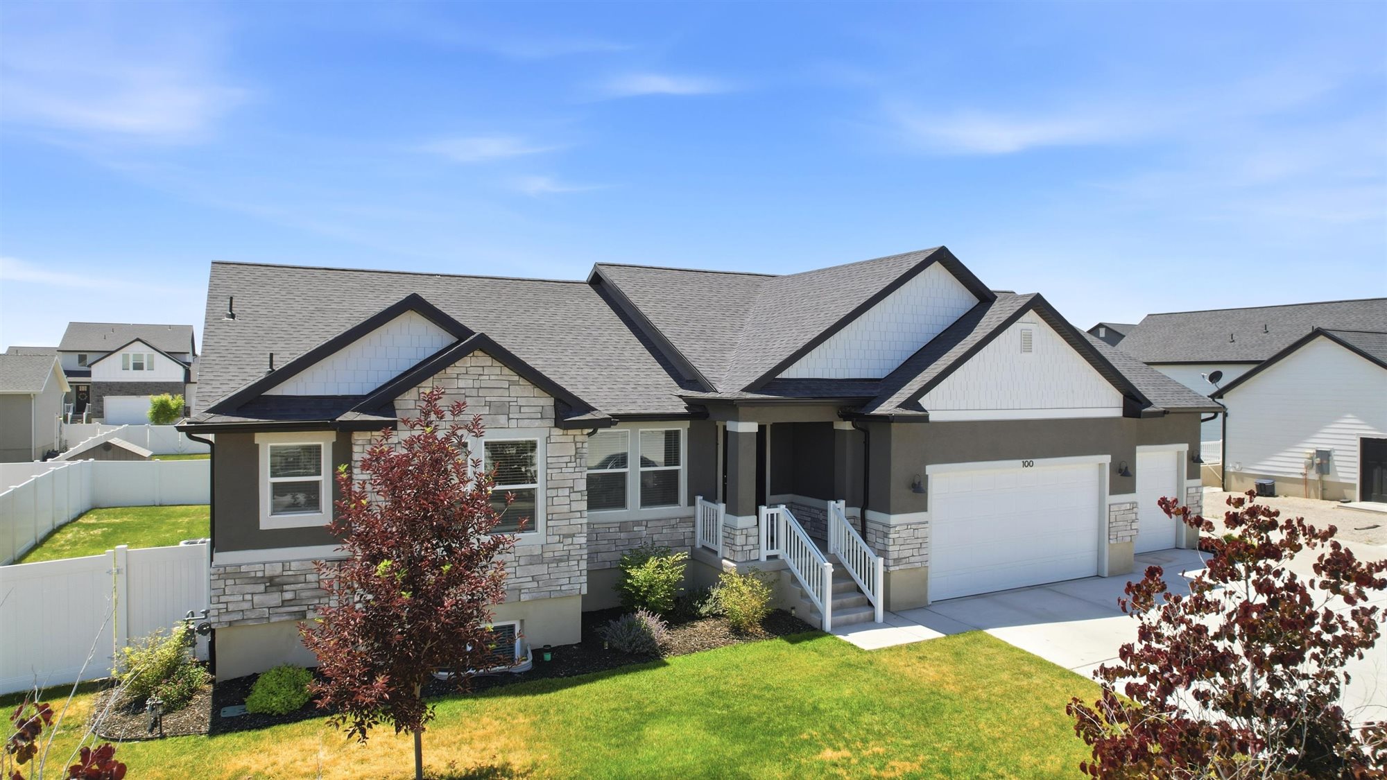 Exterior featuring a dark gray stucco finish with light-colored stone accents, a multi-gabled dark shingle roof, a two-car garage, and a white picket fence enclosing the backyard